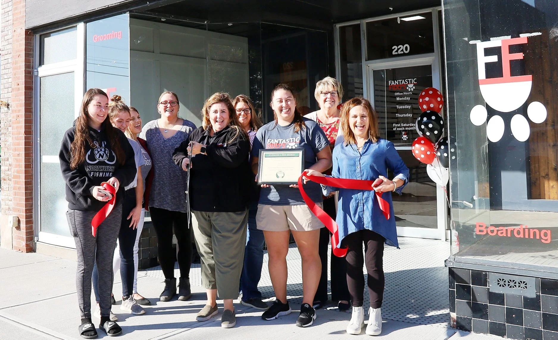 Group of women and girls standing outside a storefront for a ribbon-cutting ceremony, with one holding a large red ribbon and another holding a certificate, smiling. The storefront has balloons and signs for grooming and boarding services.
