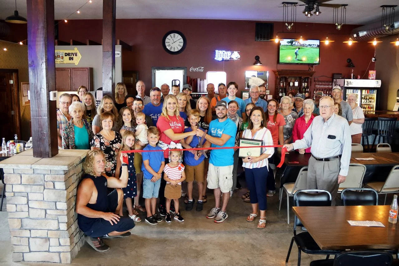 Group of people gathered for a ribbon-cutting ceremony in a restaurant or pub, with a man and woman holding scissors, surrounded by adults and children smiling.