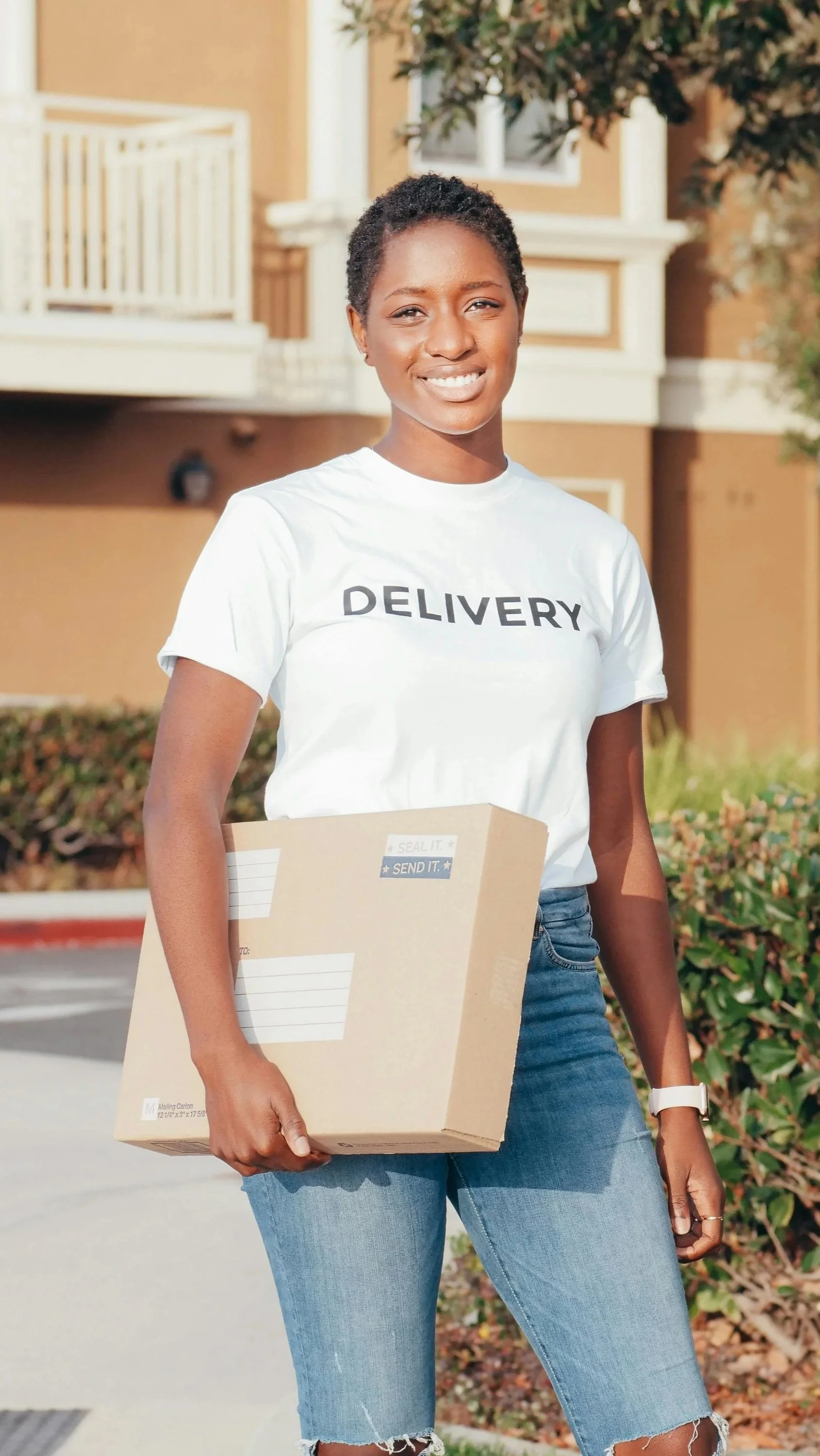 A smiling woman in a white T-shirt with 'DELIVERY' printed on it, holding a cardboard box, standing outdoors in front of a residential building in Antigua & Barbuda delivering a package