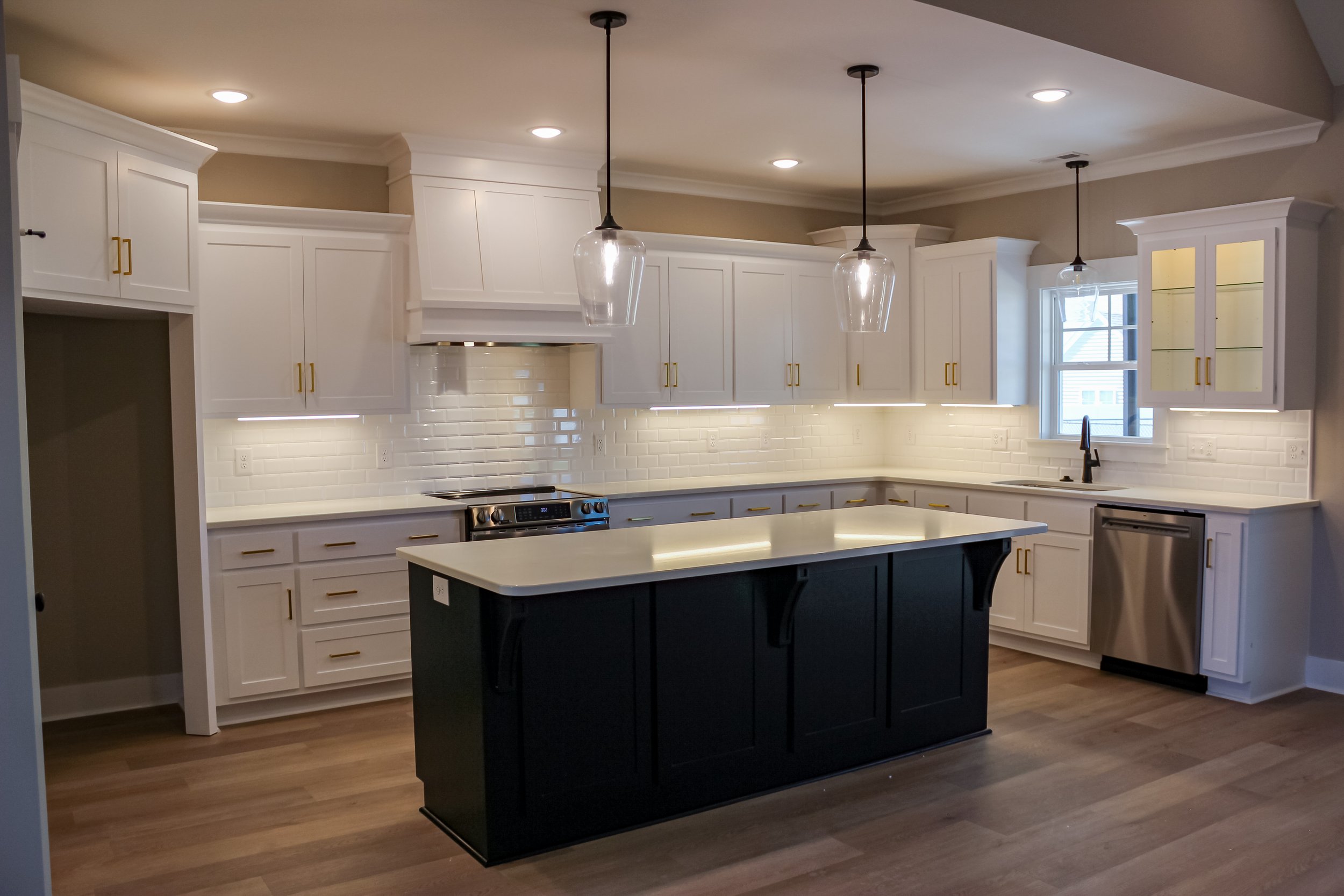 Modern kitchen with white cabinets, a black island, and hardwood floors. There are three pendant lights above the island, a window behind the sink, and a backsplash of white subway tiles.