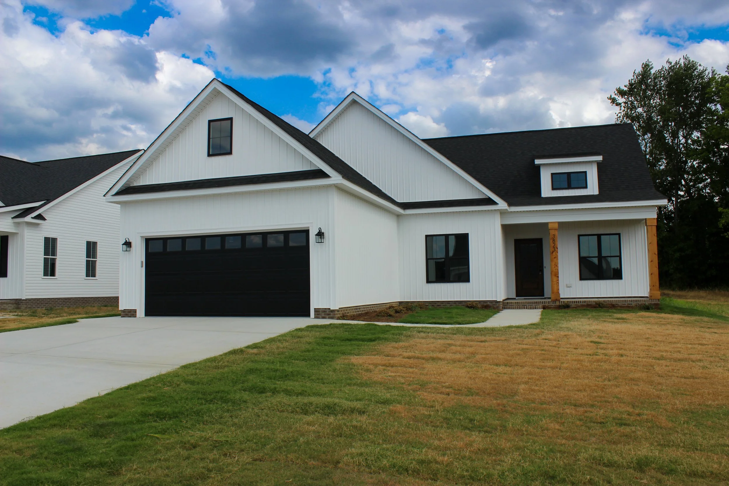 Newly constructed white house with black garage door, black window frames, and black front door, with some wooden accents around the porch, on a freshly mowed lawn under a partly cloudy sky.