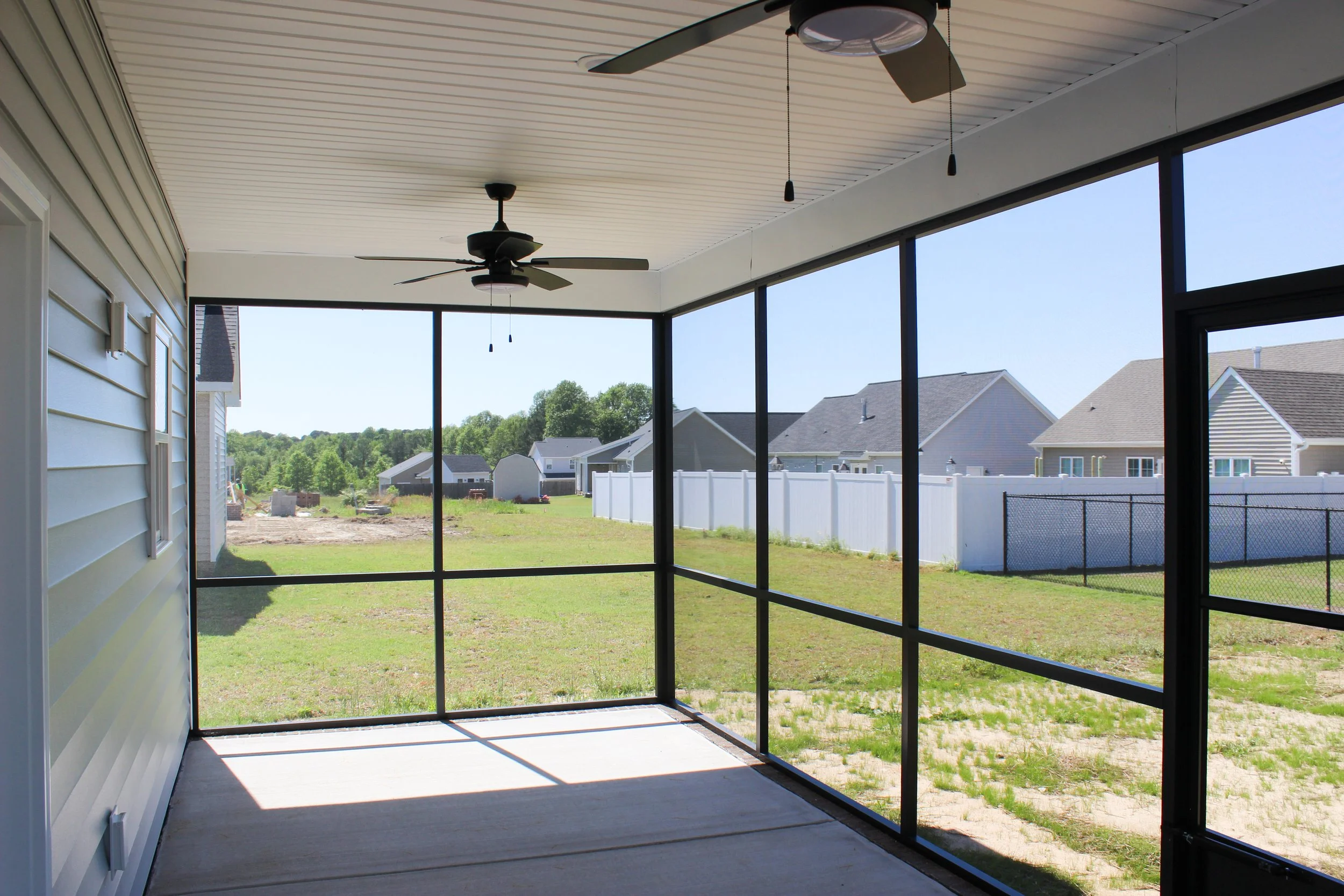 View of a screened-in porch with ceiling fans overlooking a backyard with grass, a white fence, and neighboring houses under a clear blue sky.