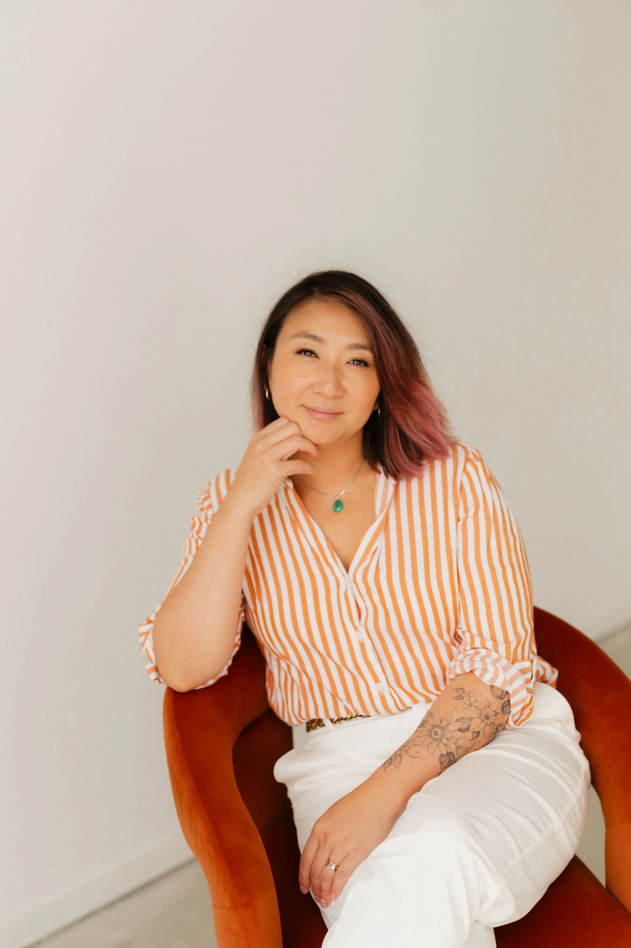 A woman sitting in an orange chair against a plain white wall, wearing a vertically striped orange and white blouse, white pants, and a green pendant necklace. She has shoulder-length hair with pink highlights and tattoos on her forearm, and she is smiling with her hand resting on her chin.