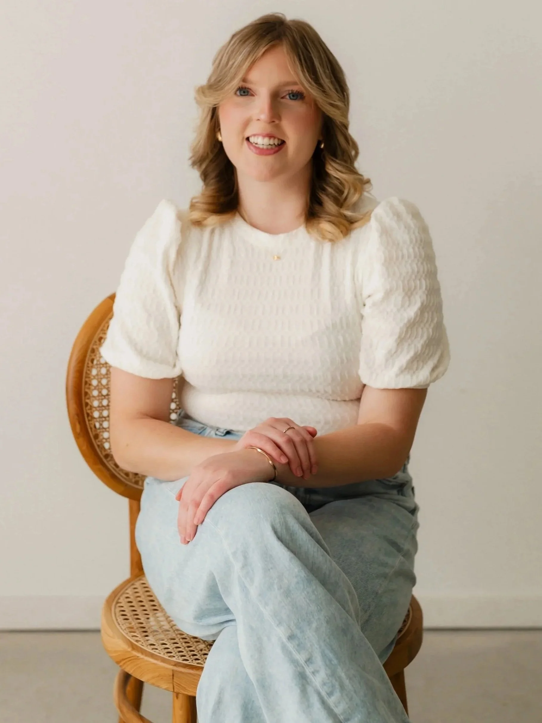 A young woman with wavy blonde hair, wearing a white textured top and light blue jeans, sitting on a wooden chair with a cane backrest, smiling at the camera against a plain white background.