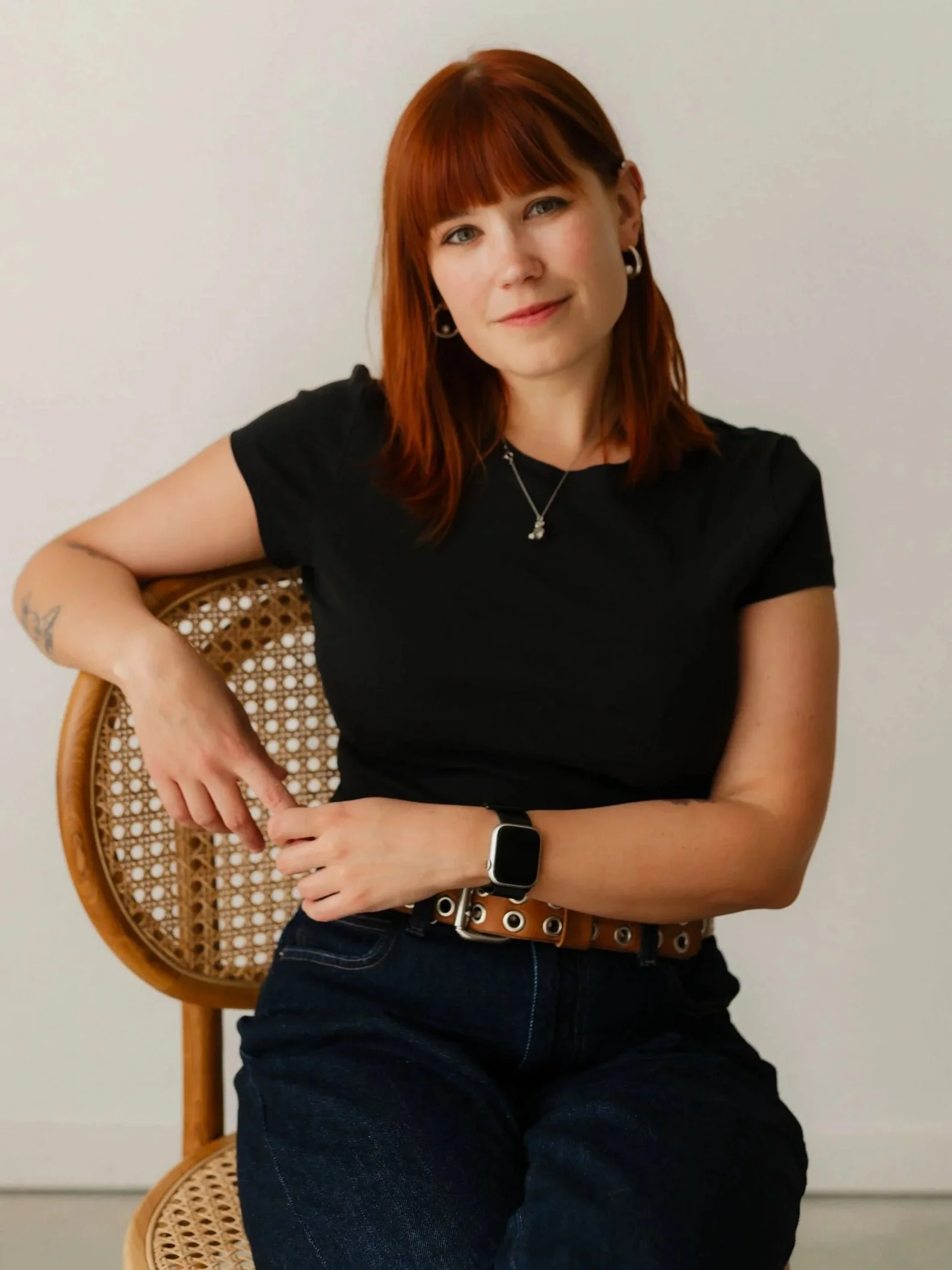 A woman with red hair, wearing a black t-shirt, jeans, and a smartwatch, sitting on a cane-backed chair with woven rattan detailing, posing with her arm resting on the chair back in front of a plain white wall.