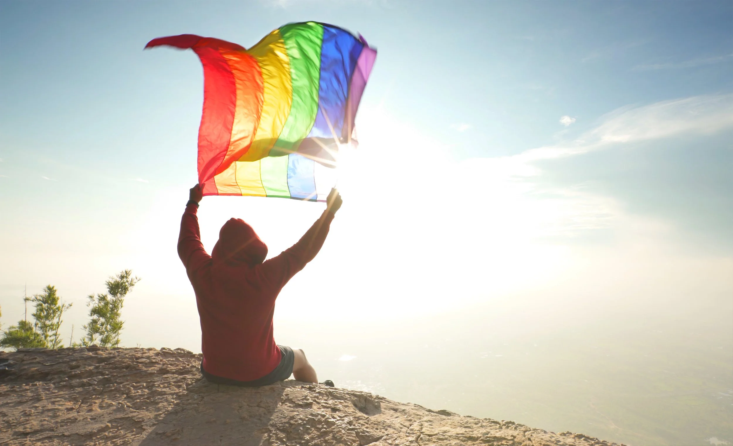 Person sitting on a rocky surface holding a rainbow flag with the sun shining brightly behind them, clear sky in the background.