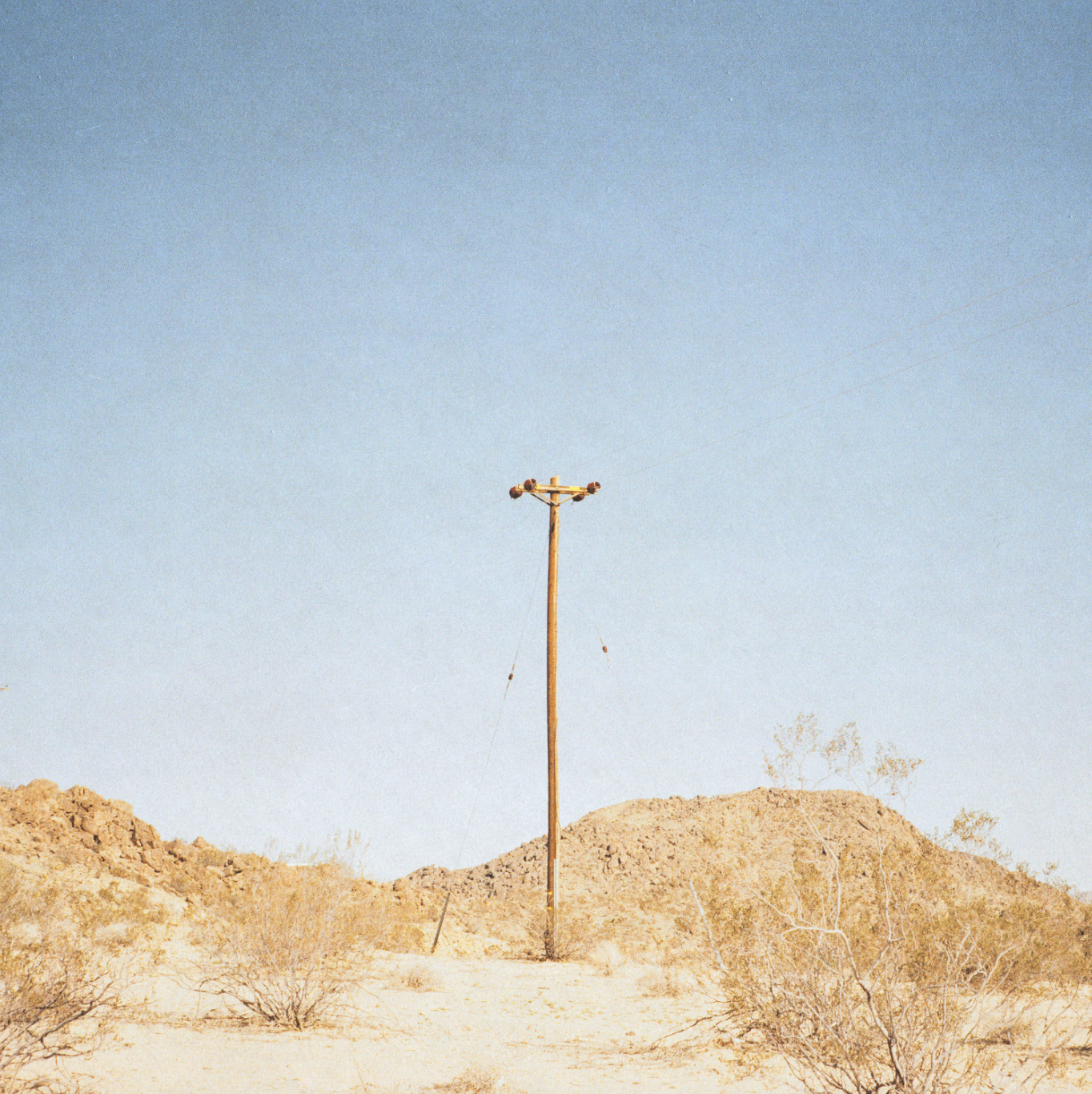 A lone wooden utility pole stands in a desert landscape with small bushes and rocky hills, under a clear blue sky.