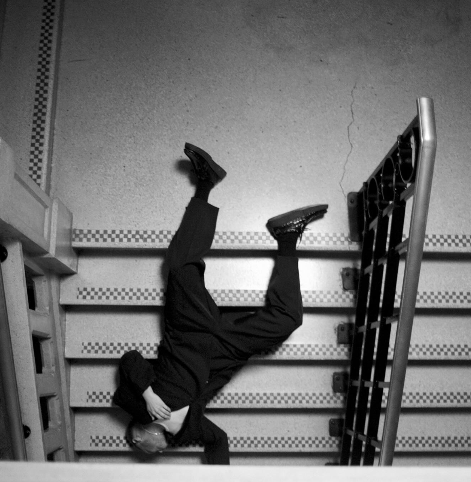 A person lying on a staircase with a hand on their face and the body partially hanging off the stairs, wearing dark clothing and sneakers.