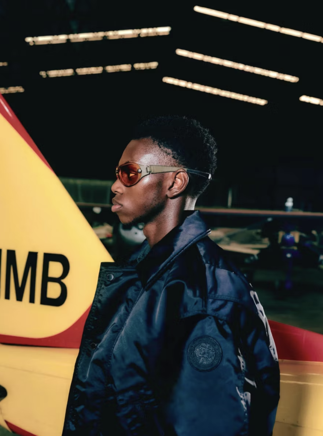 A young man wearing sunglasses and a dark jacket stands in front of a yellow airplane with a large black letter 'M' on it, inside an indoor hangar with dim lighting.