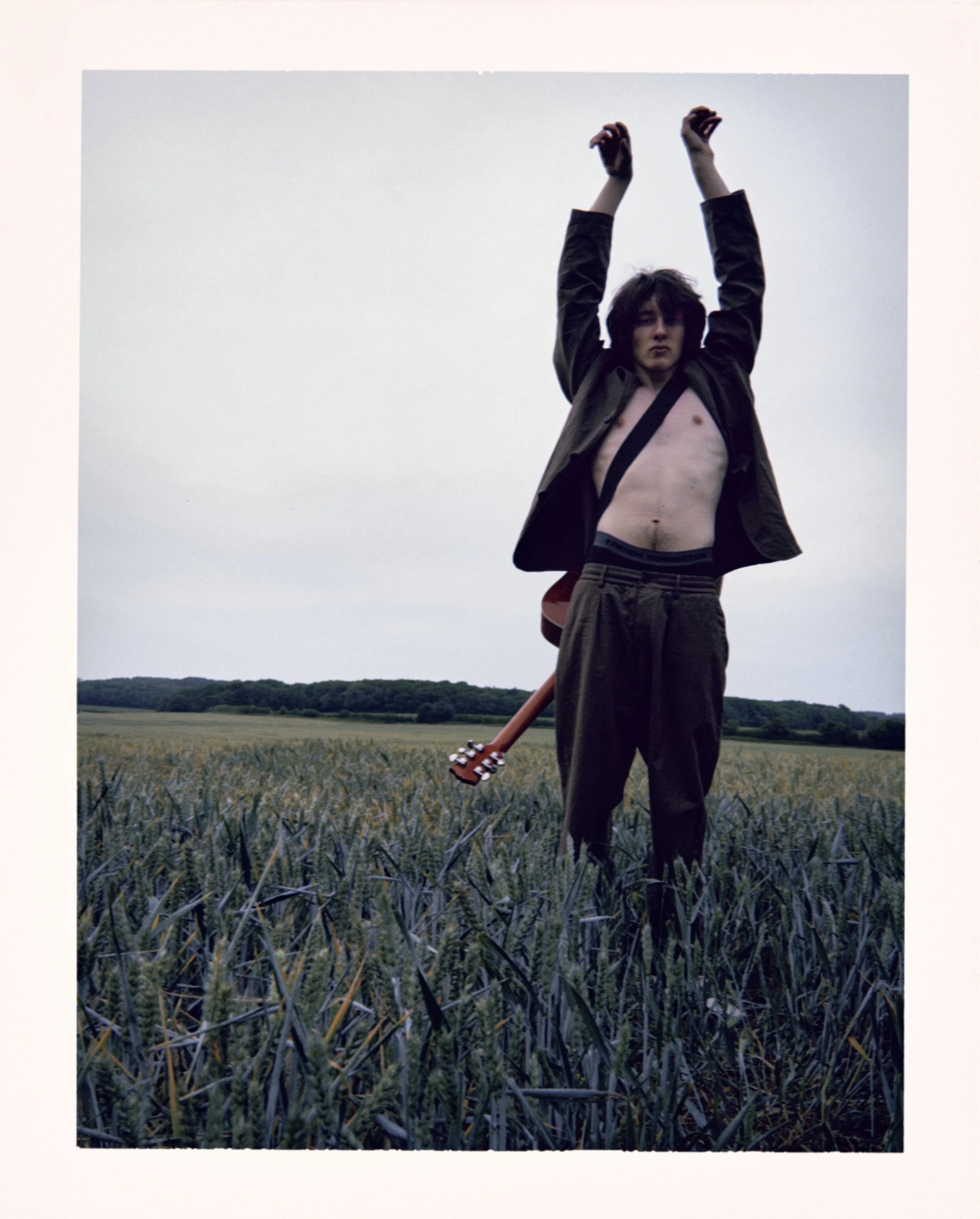 A young man standing topless in a field of tall grass or wheat, wearing loose pants and an open jacket, with an electric guitar slung over his shoulder, raising his arms above his head against a cloudy sky.
