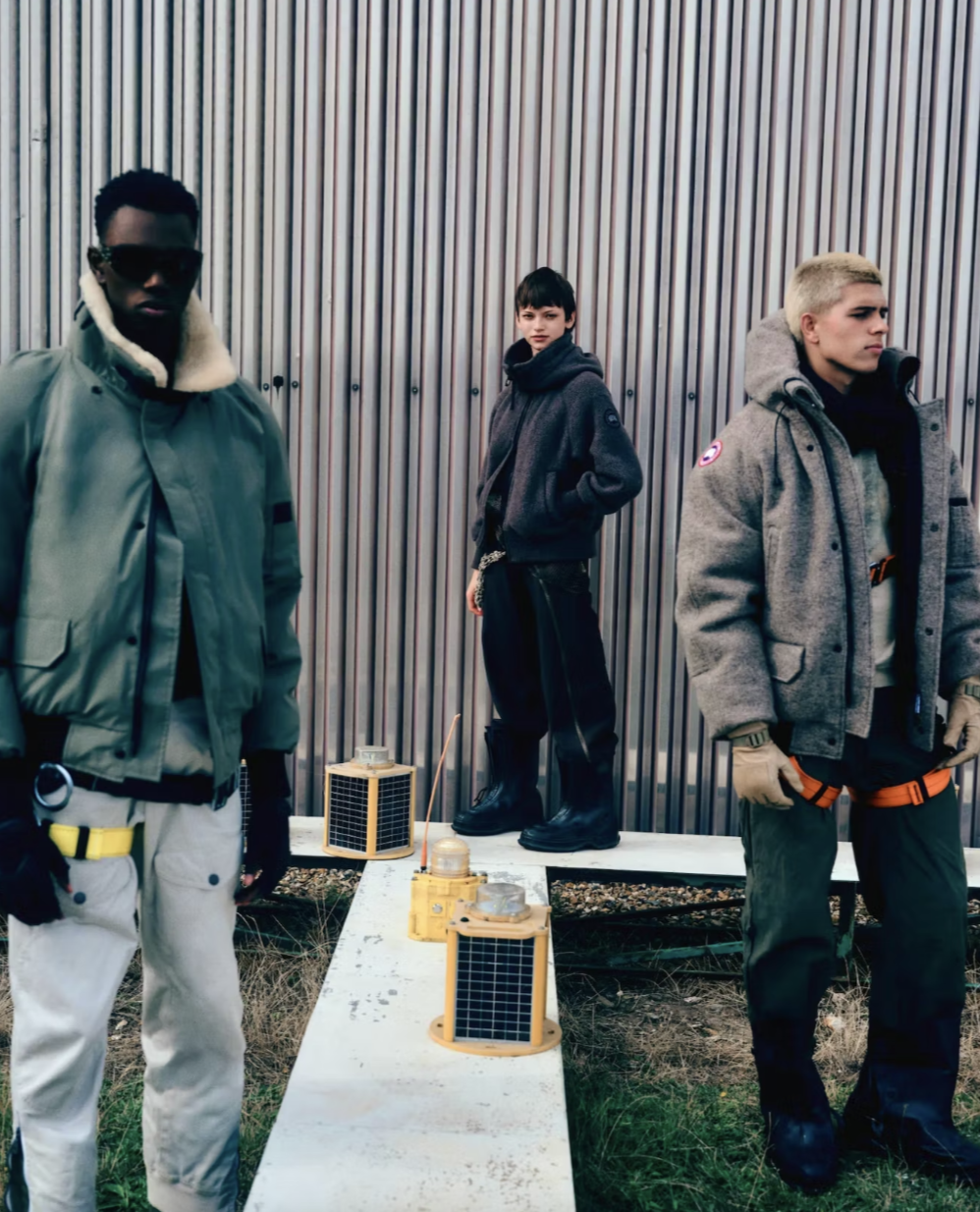 Three young men standing outdoors in front of a metal wall with small solar-powered lanterns on a concrete platform.