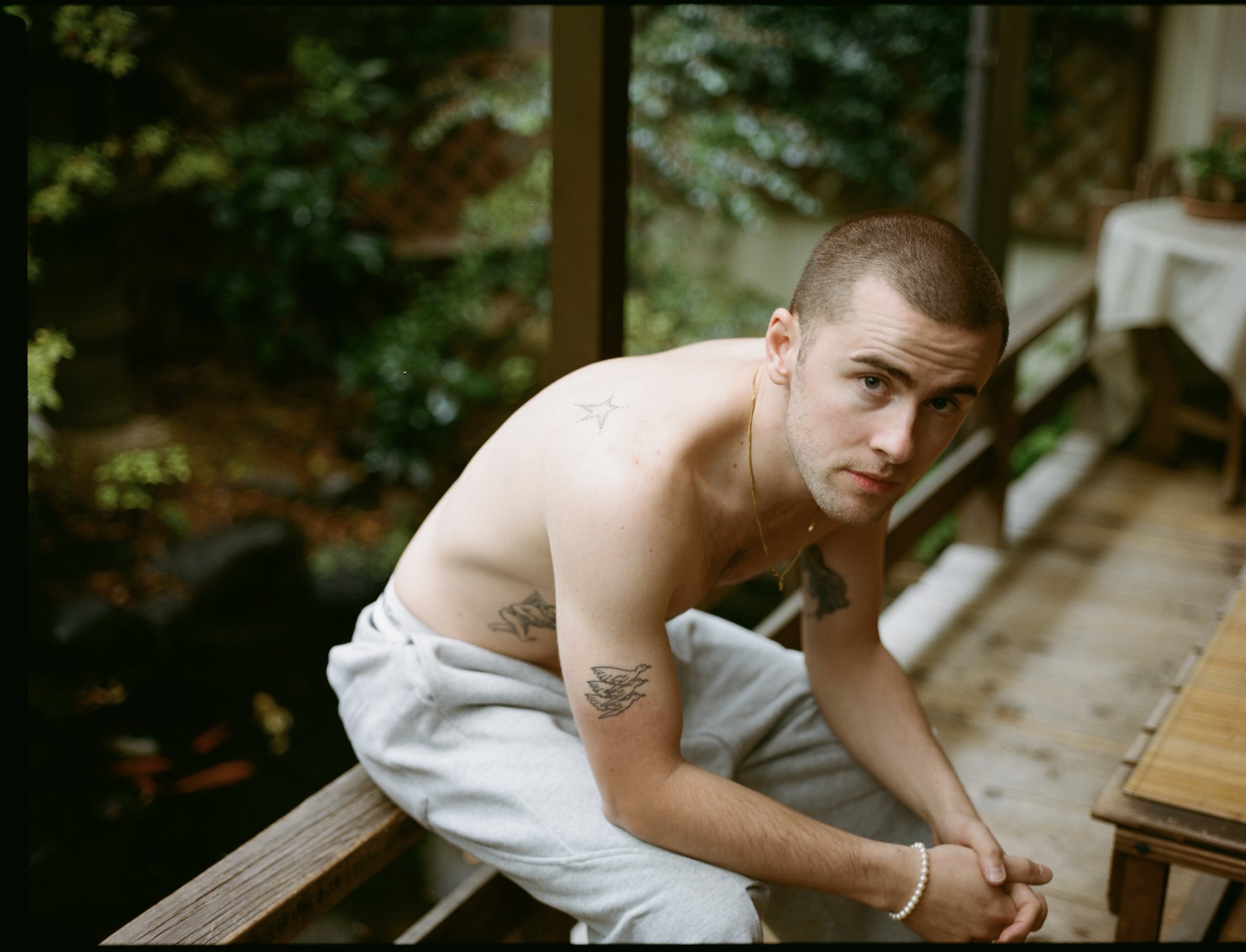 A shirtless young man with short hair and tattoos on his arms and chest, sitting on a wooden deck. He's looking at the camera with a serious expression, outdoors with greenery and garden furniture in the background.