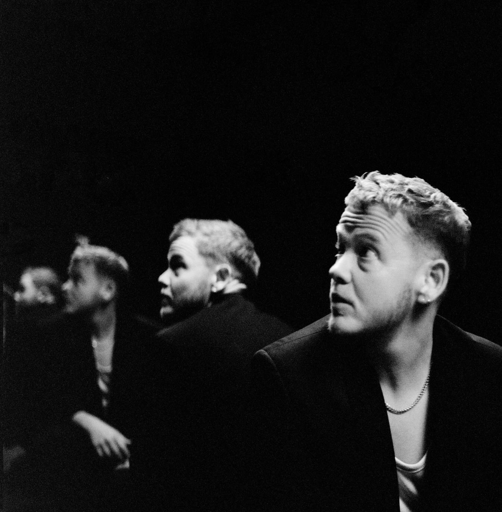 A black and white photo of four young men sitting in a row, with the focus on the man on the far right who is looking to his left, while the others look in different directions, set against a dark background.