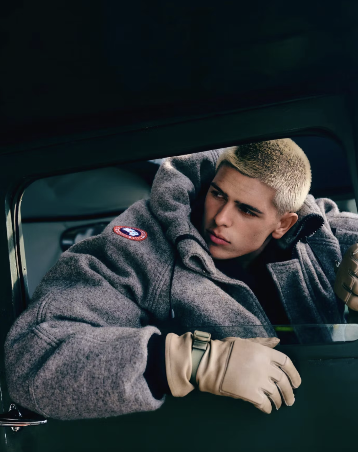A young man with platinum blonde hair wearing a gray jacket and tan gloves, looking out from a military vehicle window.