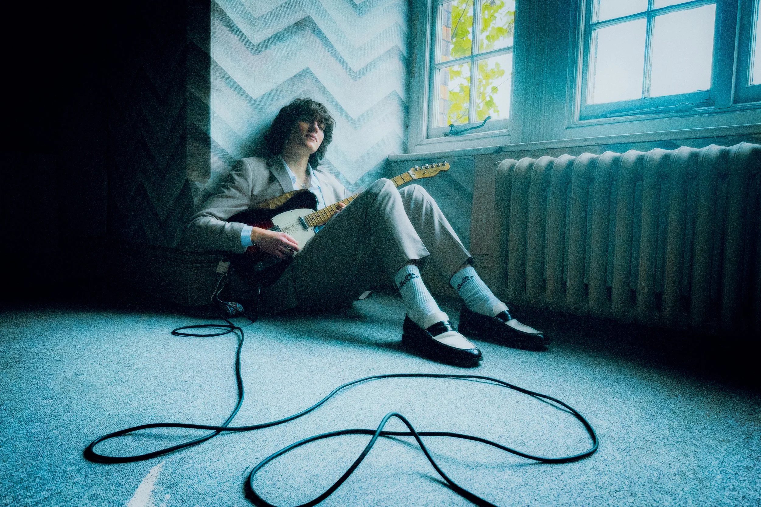 Person sitting on the floor, playing an electric guitar near a window with daylight, wearing a light-colored suit, with curly hair, and positioned against a chevron-patterned wall.