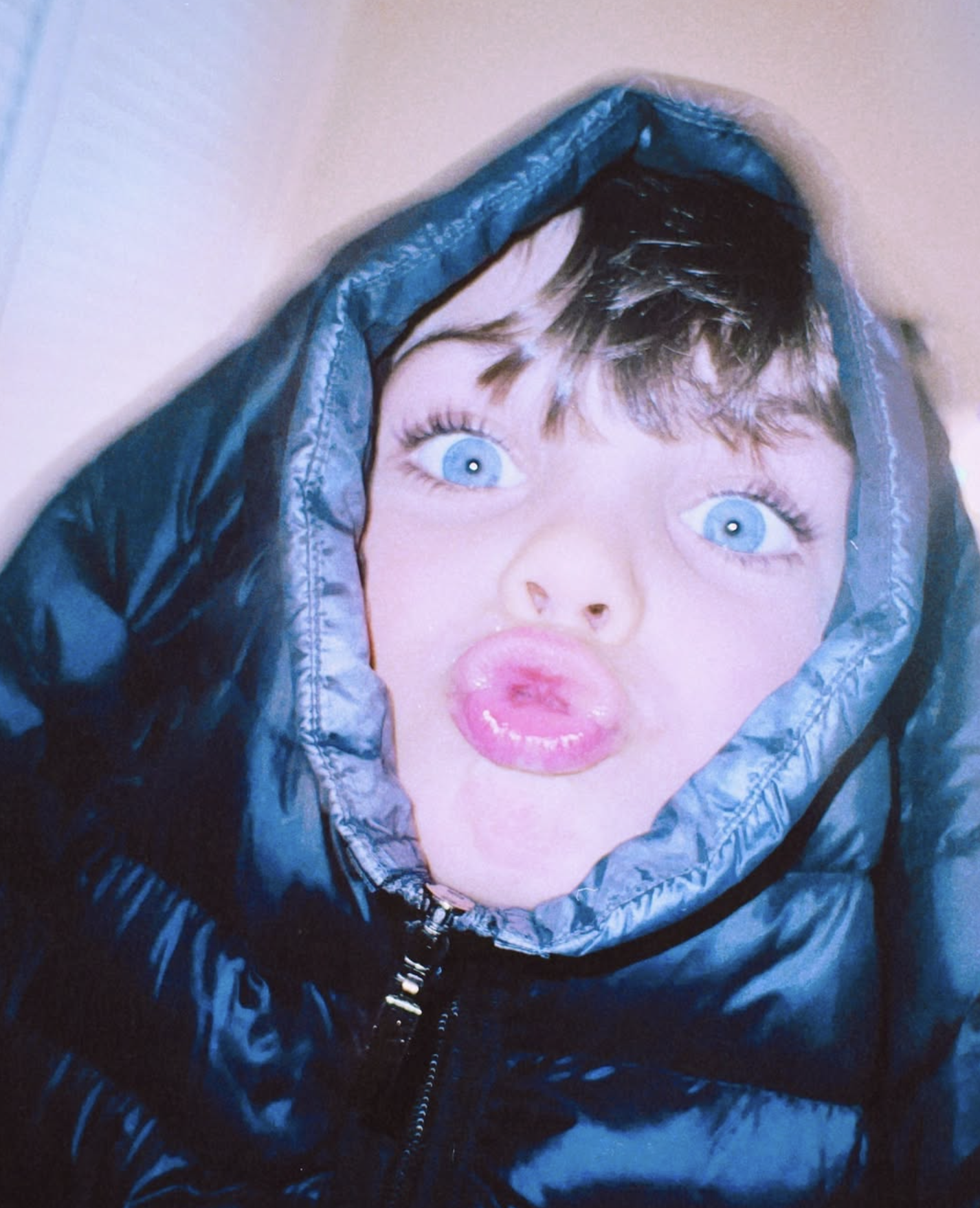Woman with blue eyes and dark hair making a pout face, wearing a dark blue puffer jacket with the hood over her head.