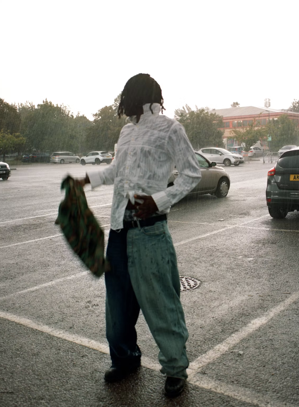 A person with dreadlocks wearing a white jacket and baggy jeans stands in a parking lot during heavy rain, holding a colorful cloth in their hand.