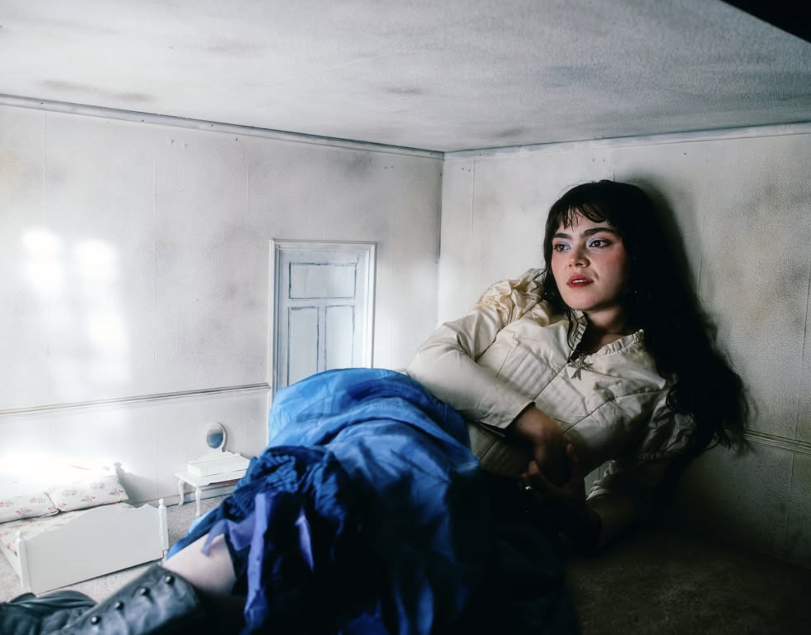 A woman with long dark hair and makeup, wearing a beige jacket, lying on her side in a small, plain room with white walls, a bed with floral sheets, and a small white nightstand.