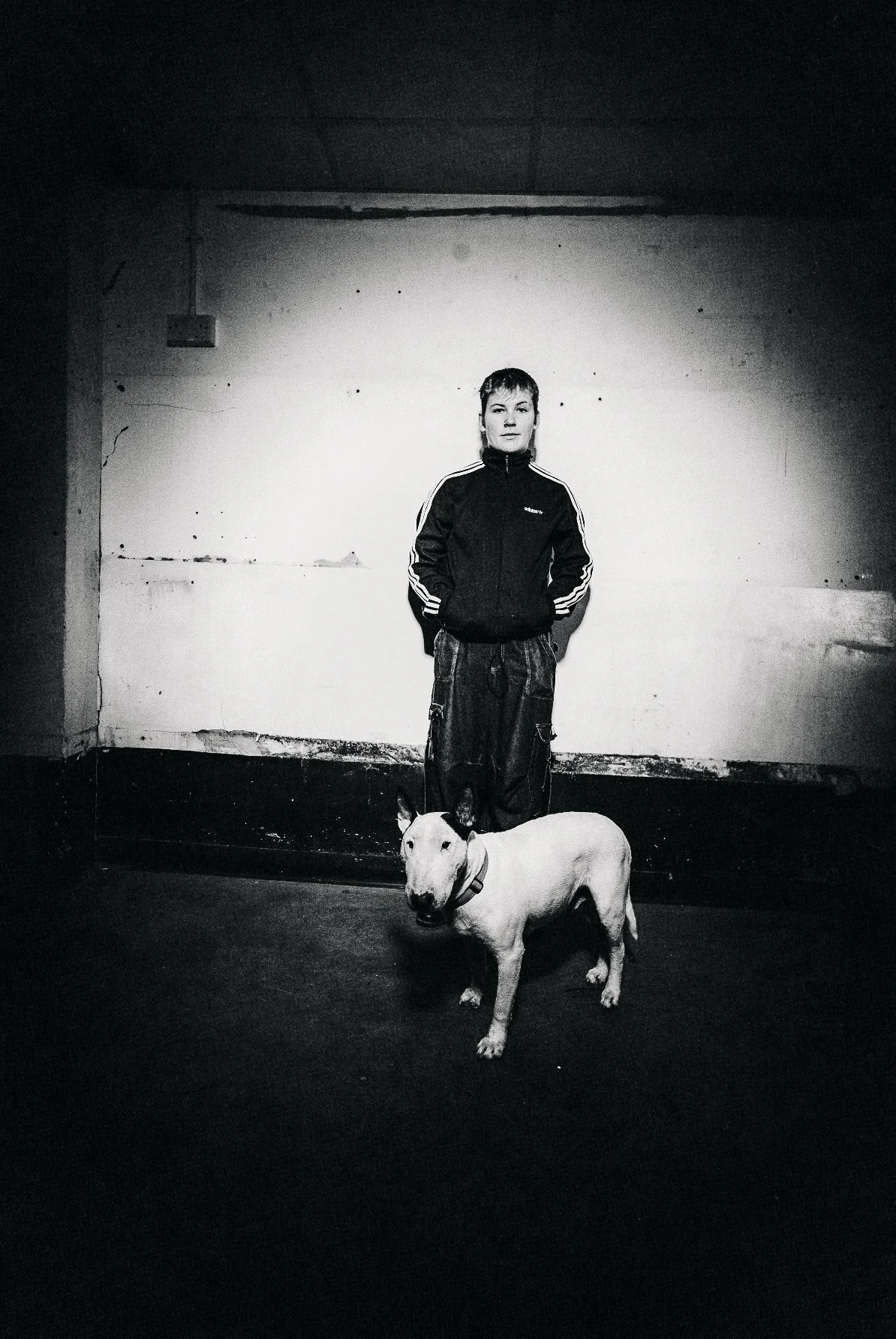 A woman in a black athletic jacket with white stripes and baggy pants stands with her hands behind her back, next to a white Bull Terrier dog with a collar, in a dimly lit, rough indoor space with peeling walls.