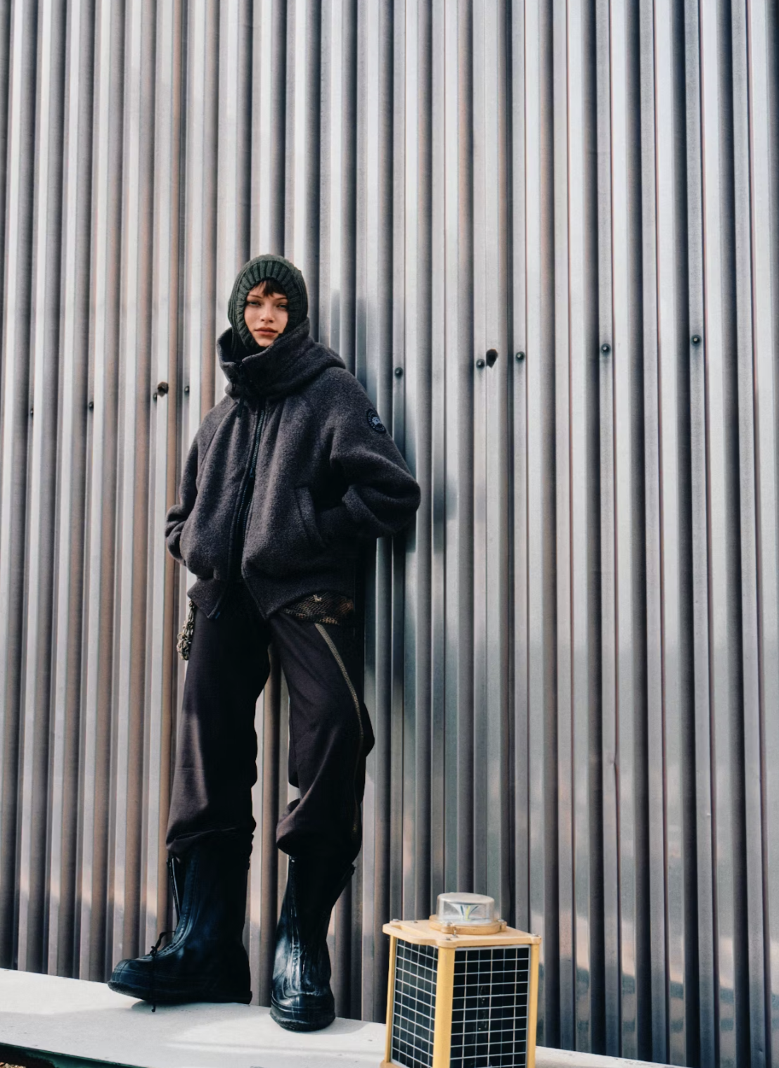 A person standing against a metallic corrugated wall, wearing a dark jacket, pants, boots, and a knit hat, with hands in pockets and a small yellow and black grid-patterned object on the ground nearby.