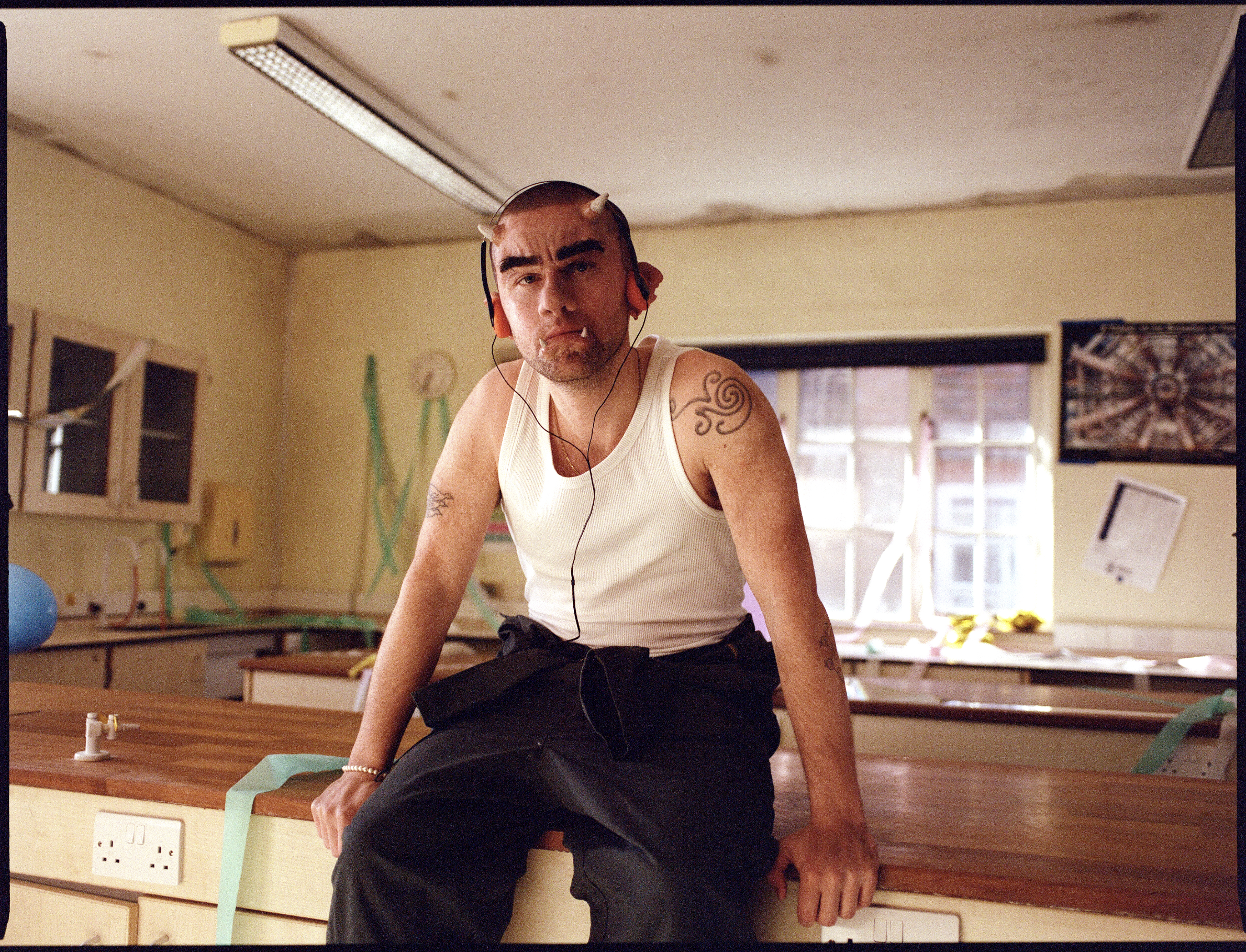 A young man with horns on his head, tattoos on his arms, and fangs, wearing a white tank top and black pants, sits on a wooden counter in a room that resembles a classroom or workshop with beige walls and various items like a blue ball, paperwork, an