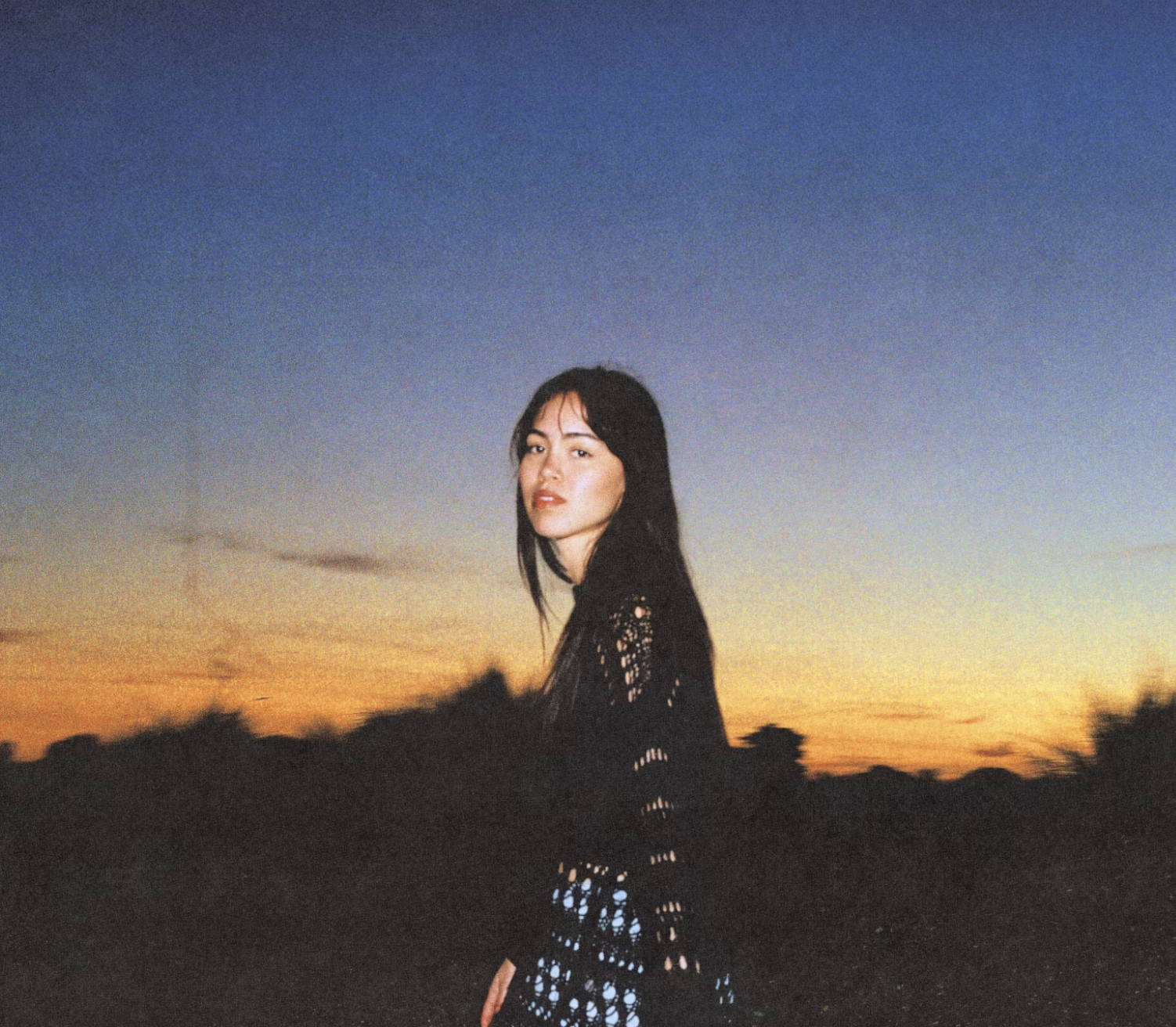 A woman with dark hair wearing a black, crochet-patterned dress stands outdoors during sunset with a colorful sky in the background.
