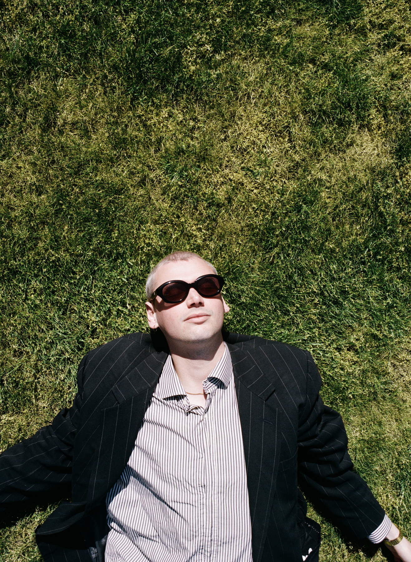 A man with blond hair, wearing large black sunglasses, a black pinstripe blazer, and a striped shirt, is lying on green grass, looking up at the sky.