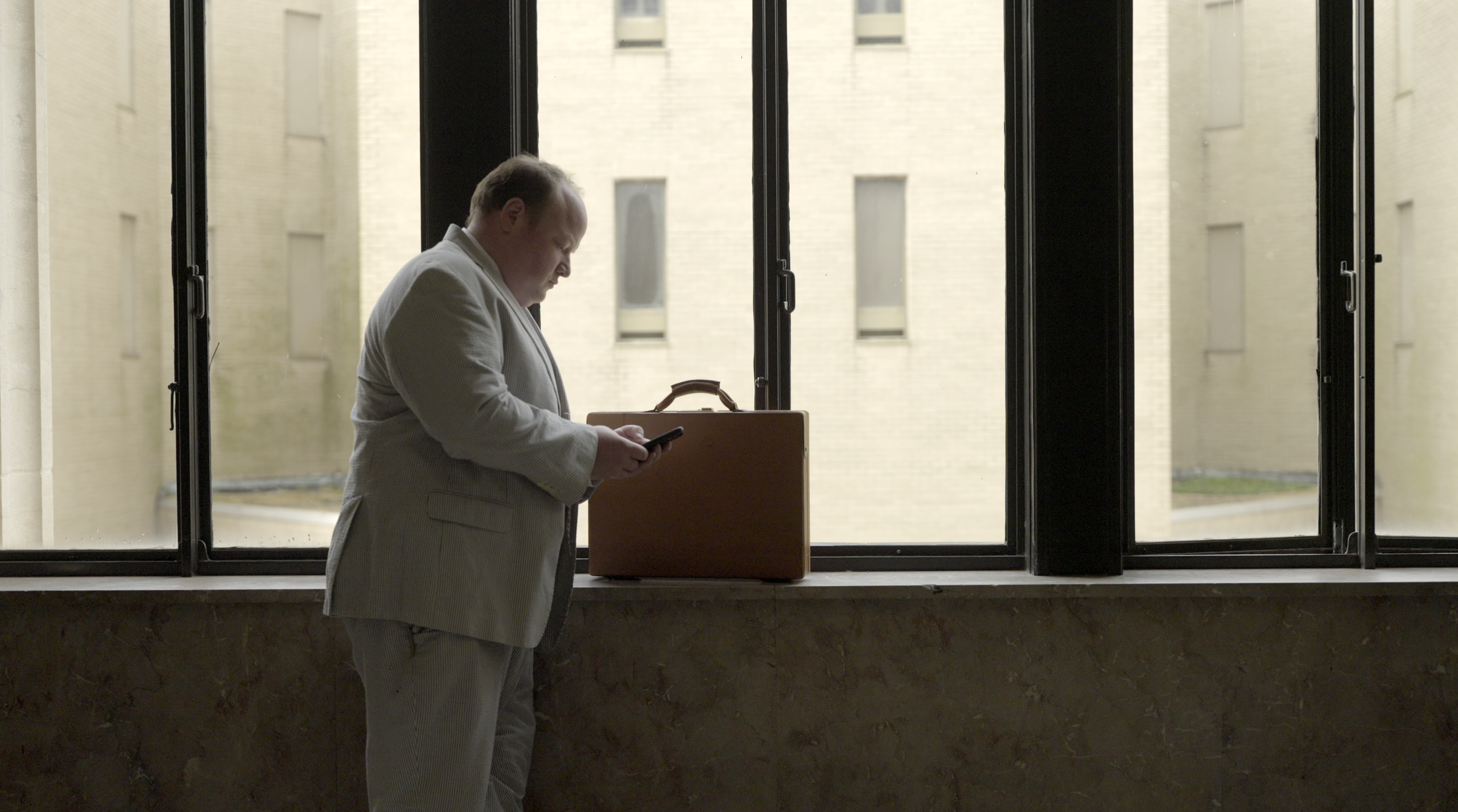 Man in a beige suit standing by a window, looking at his phone, with a brown briefcase on the windowsill.