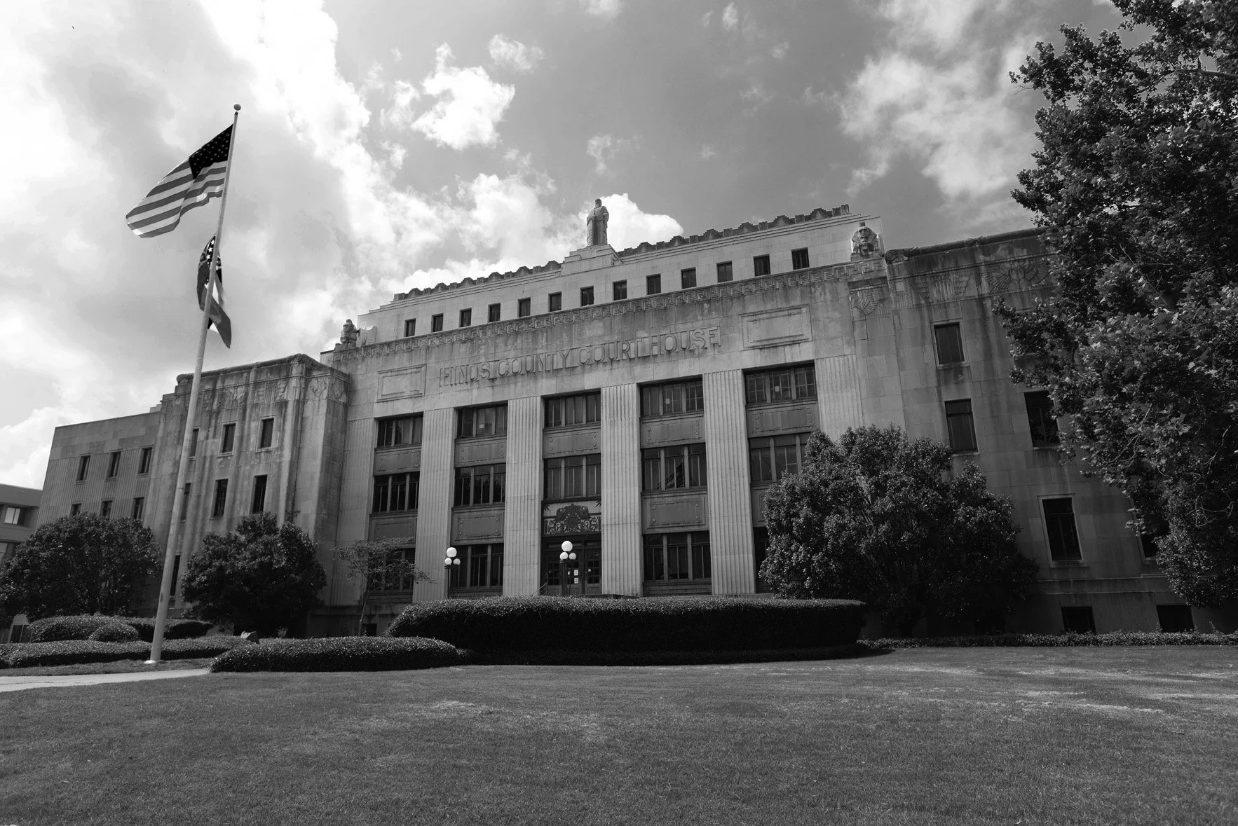 Black and white photo of the Illinois County Courthouse, a large stone building with columns at the front, American flag on display, and trees and shrubs in the foreground.