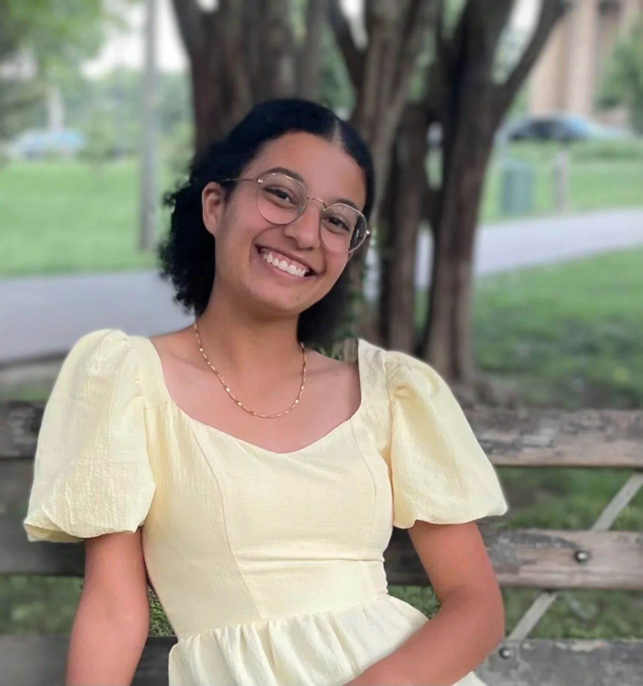 A young woman with glasses and a necklace sitting on a park bench outdoors, smiling at the camera, wearing a yellow dress with puffed sleeves.