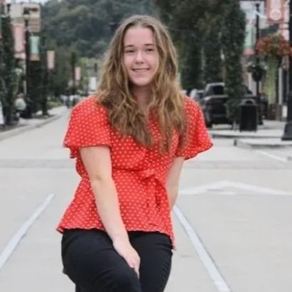 A young woman with long curly hair smiling, wearing a red polka dot blouse and black pants, kneeling outdoors on a city street.