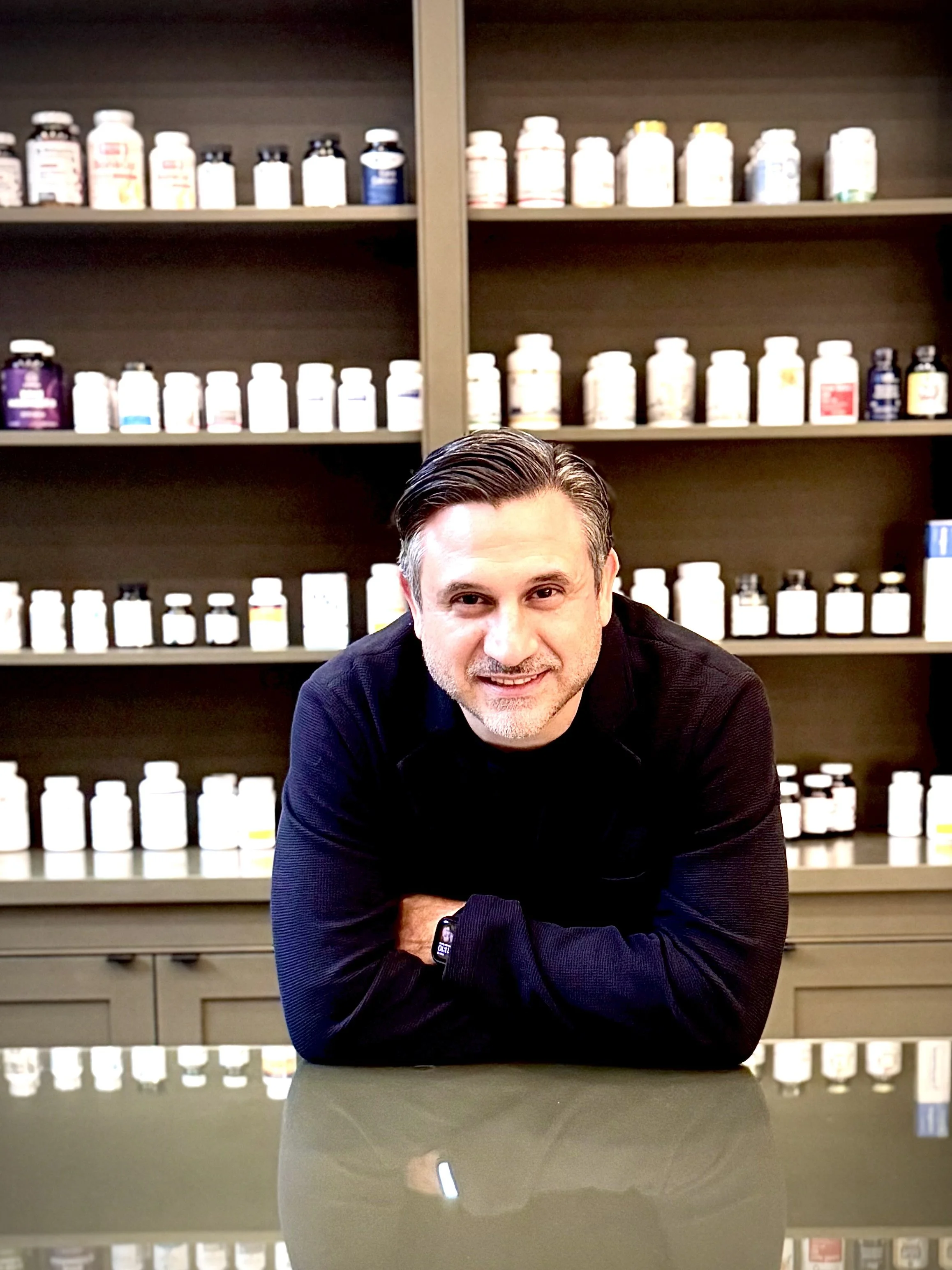 A man with dark hair and a beard smiling at a camera, leaning on a glossy table in front of shelves filled with numerous bottles of supplements or vitamins.
