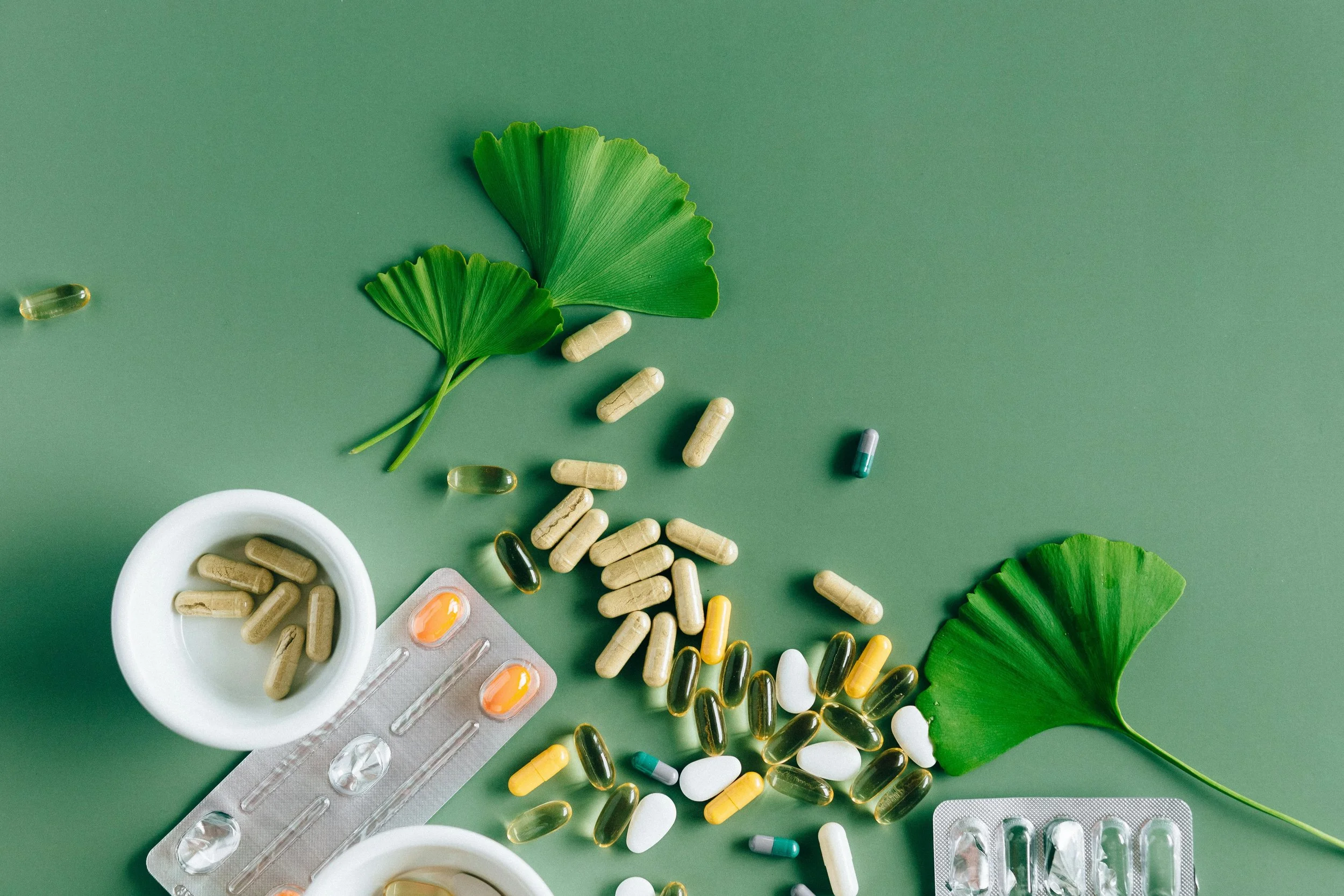 Assorted pills and capsules scattered on a green surface with two ginkgo leaves and two white bowls
