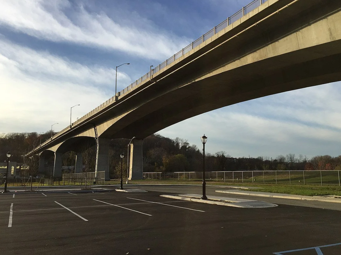 An empty parking lot with parking spaces, street lamps, and a concrete bridge overhead in a suburban area, with trees and a fence in the background.