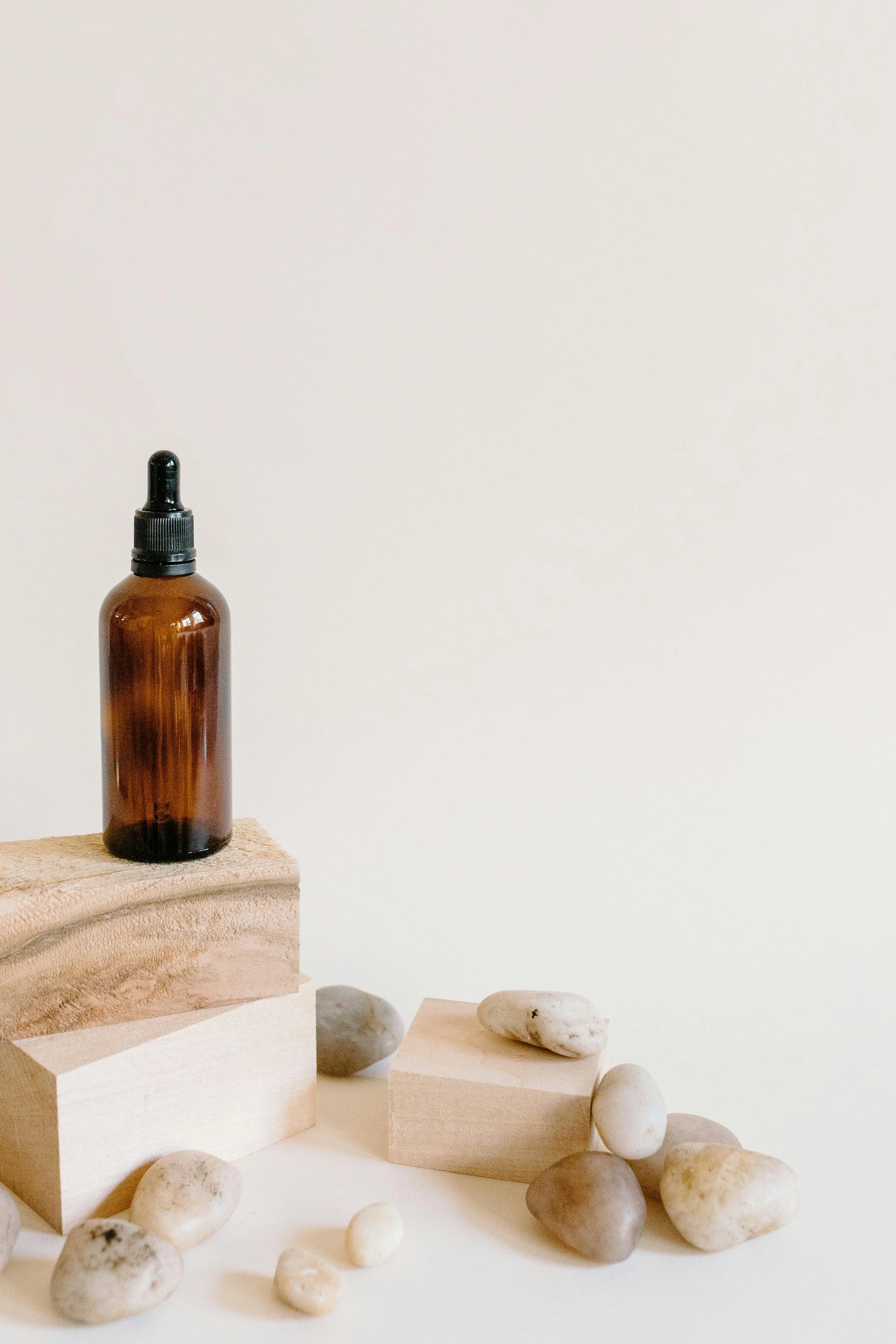 An amber glass dropper bottle on wooden blocks surrounded by smooth white stones on a white surface.