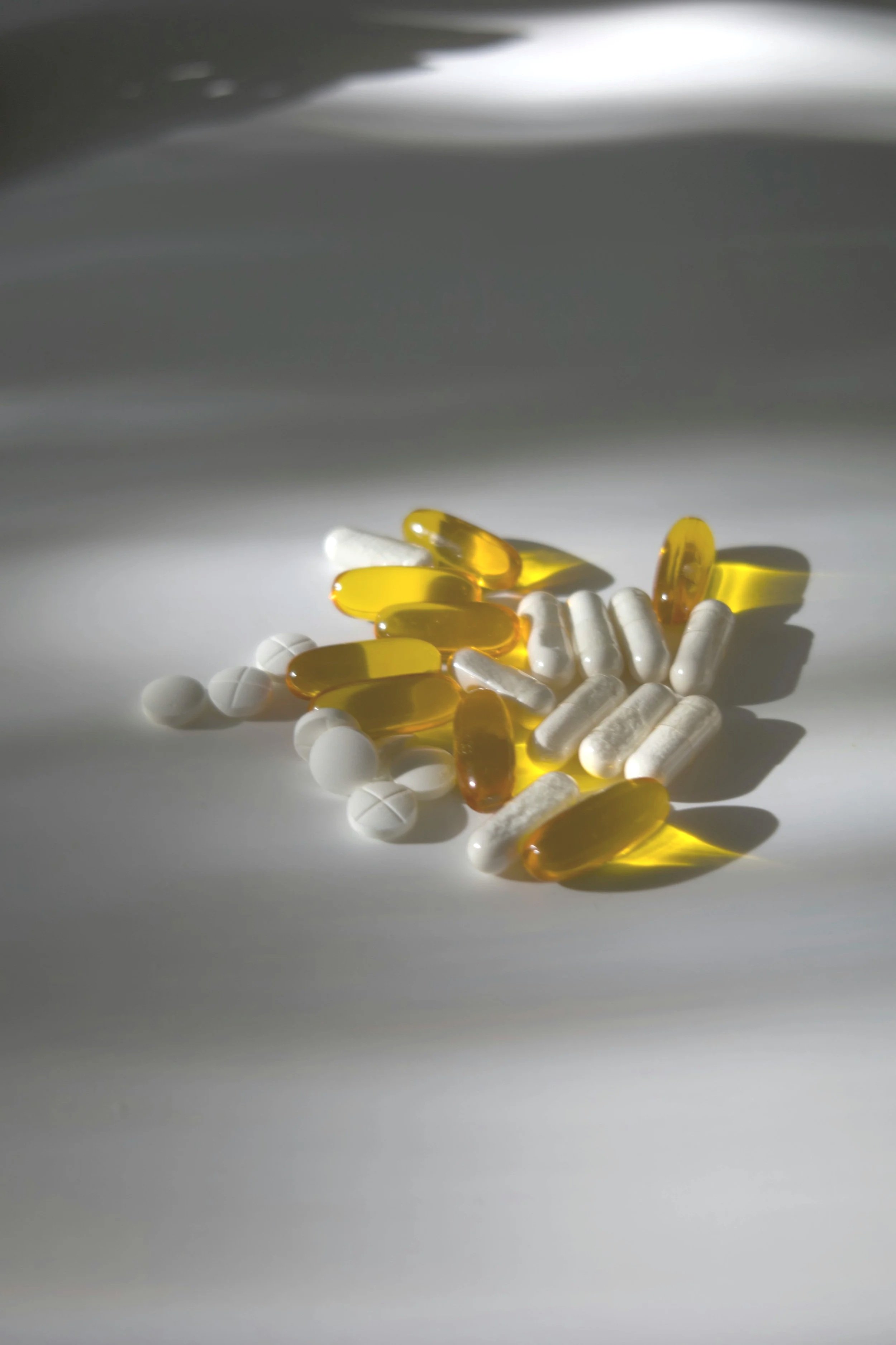A collection of various pills and capsules, including white round tablets, white oblong tablets, and yellow and translucent gel capsules, scattered on a white surface with shadows and bright light.
