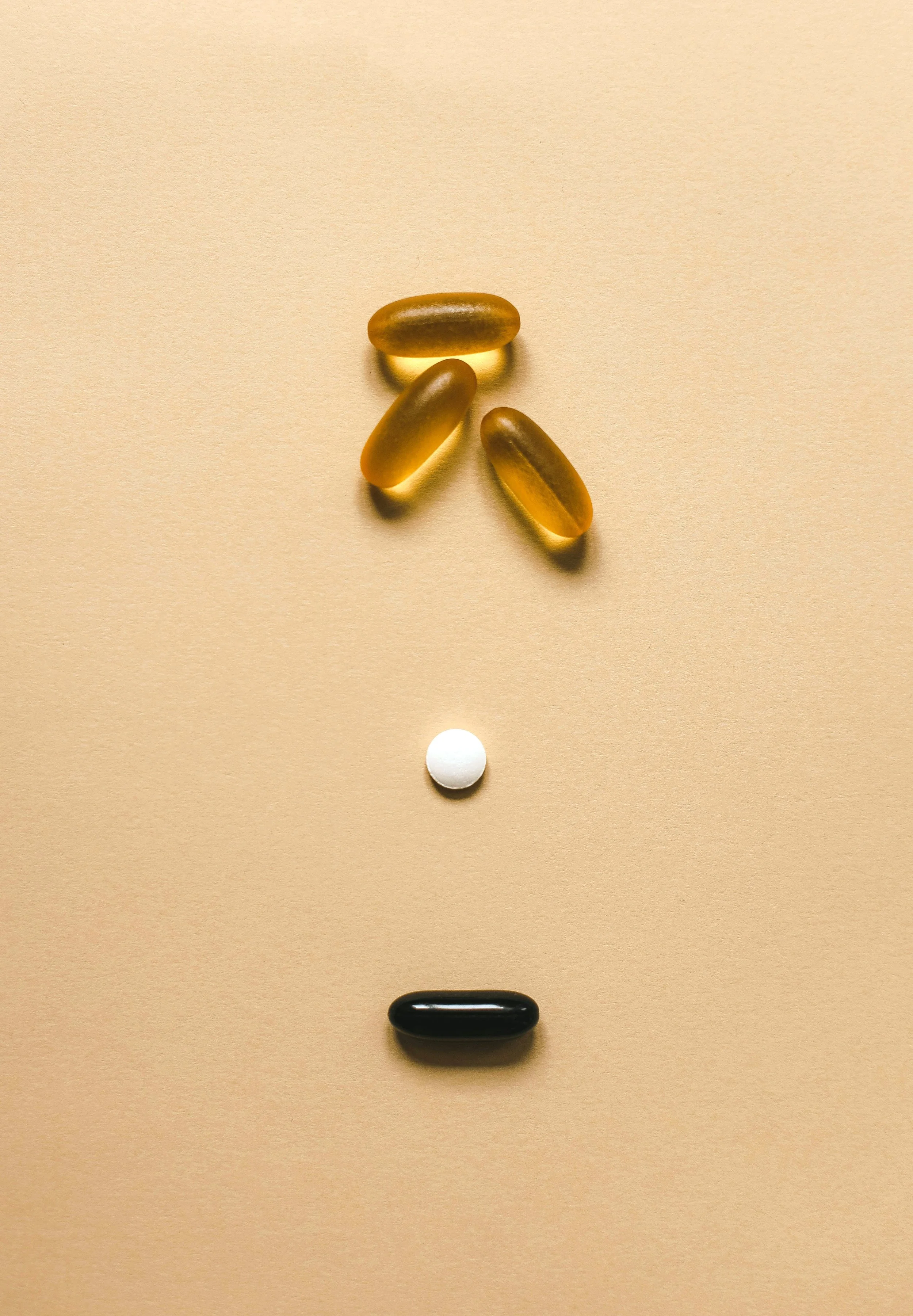 Various pills arranged on a beige background to resemble a face, with three elongated capsules at the top representing hair, a small white round pill as a nose, and a black capsule as a mouth.