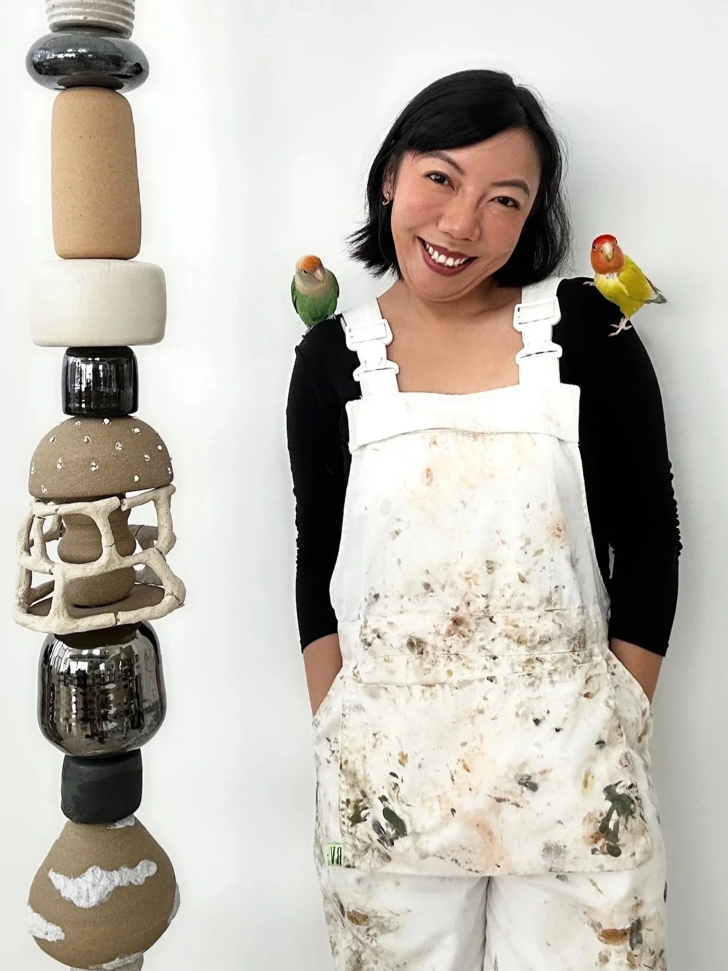 A woman with long black hair smiling, wearing a speckled sleeveless top and beige pants, standing in art gallery with abstract artwork and pottery on display.