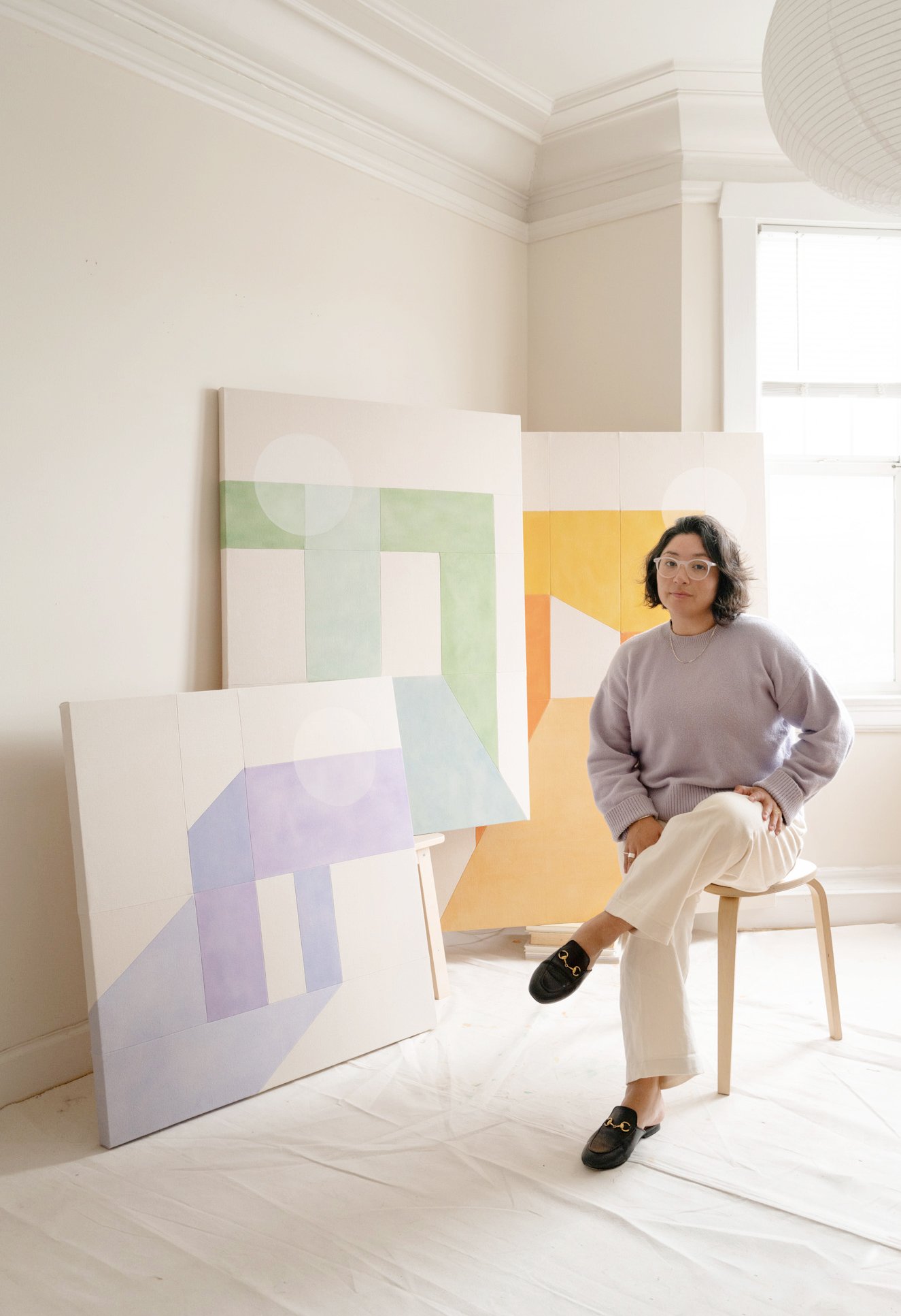 A woman with black hair, glasses, a white t-shirt, and black pants standing next to a small brown and white dog sitting on a white chair. They are indoors against a wall with four black and white modern art paintings.