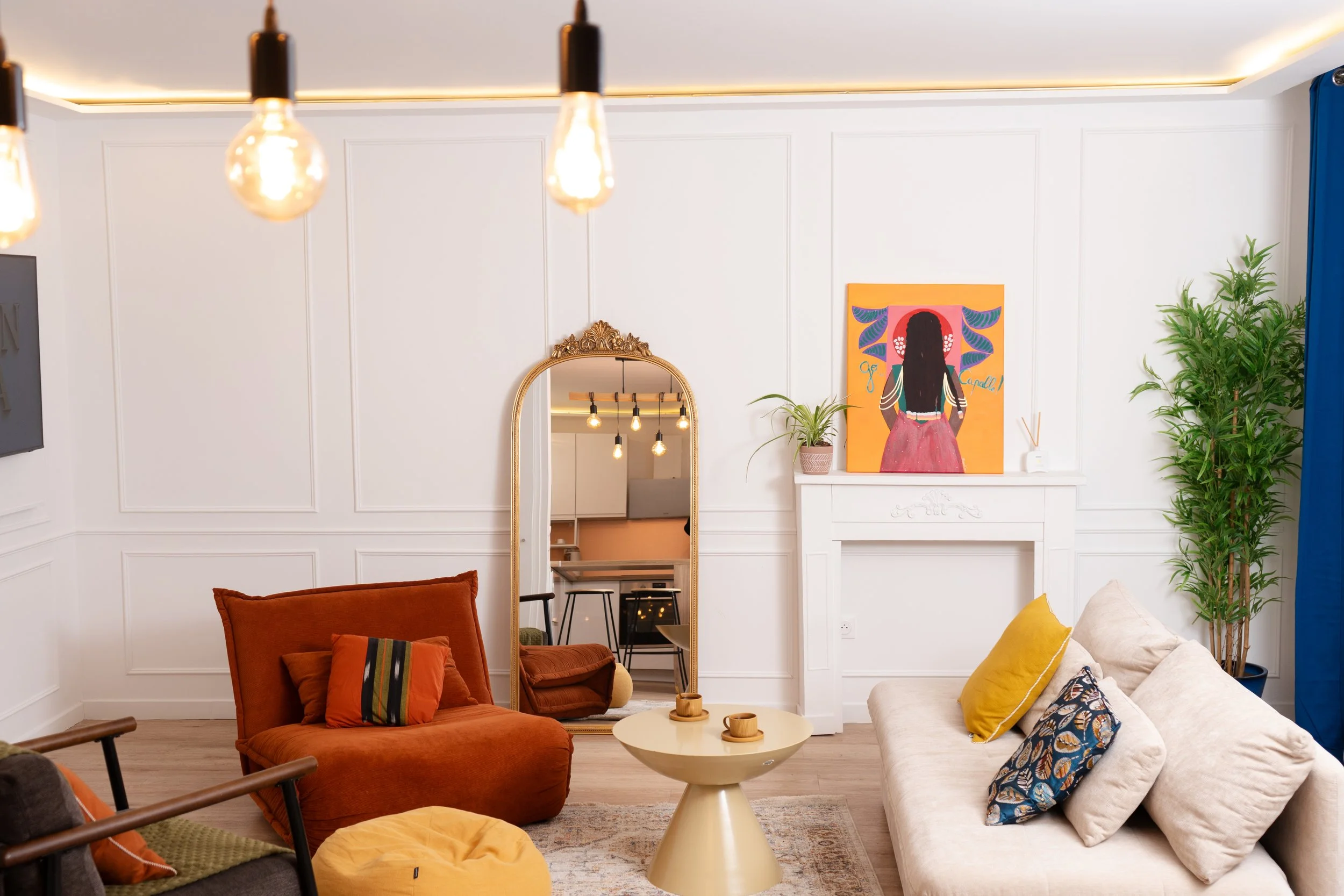 Salon moderne avec canapés beige et orange, table centrale, grand miroir doré, tableau avec silhouette féminine, plantes vertes, lampe suspendue avec ampoules visibles