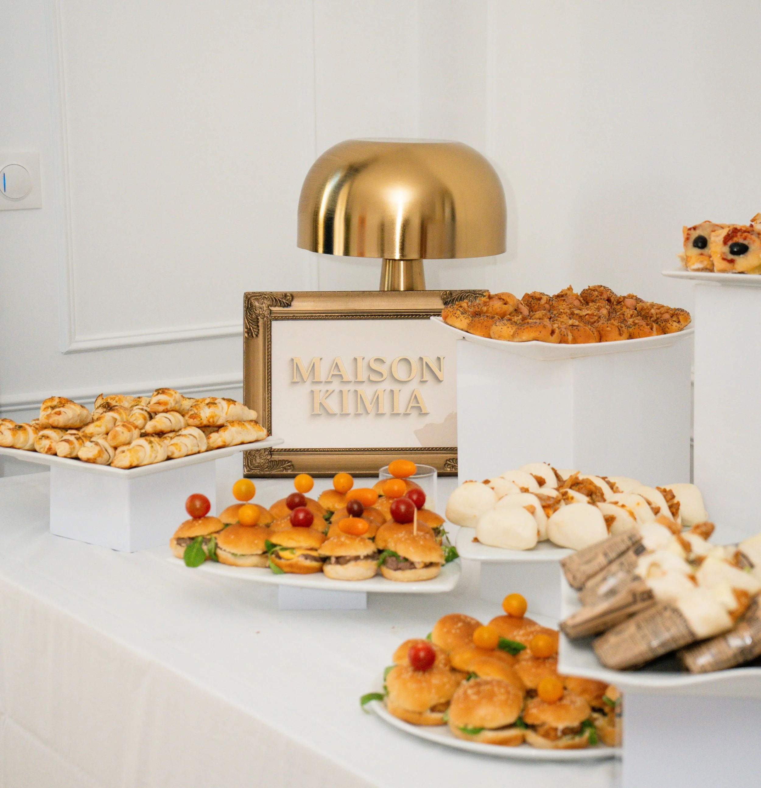 Buffet de amuse-gueules avec des petits sandwiches, des bouchées, des mini hamburgers, et des pâtisseries, avec un cadre portant l'insigne "MAISON KIMIA" et une lampe dorée en arrière-plan.