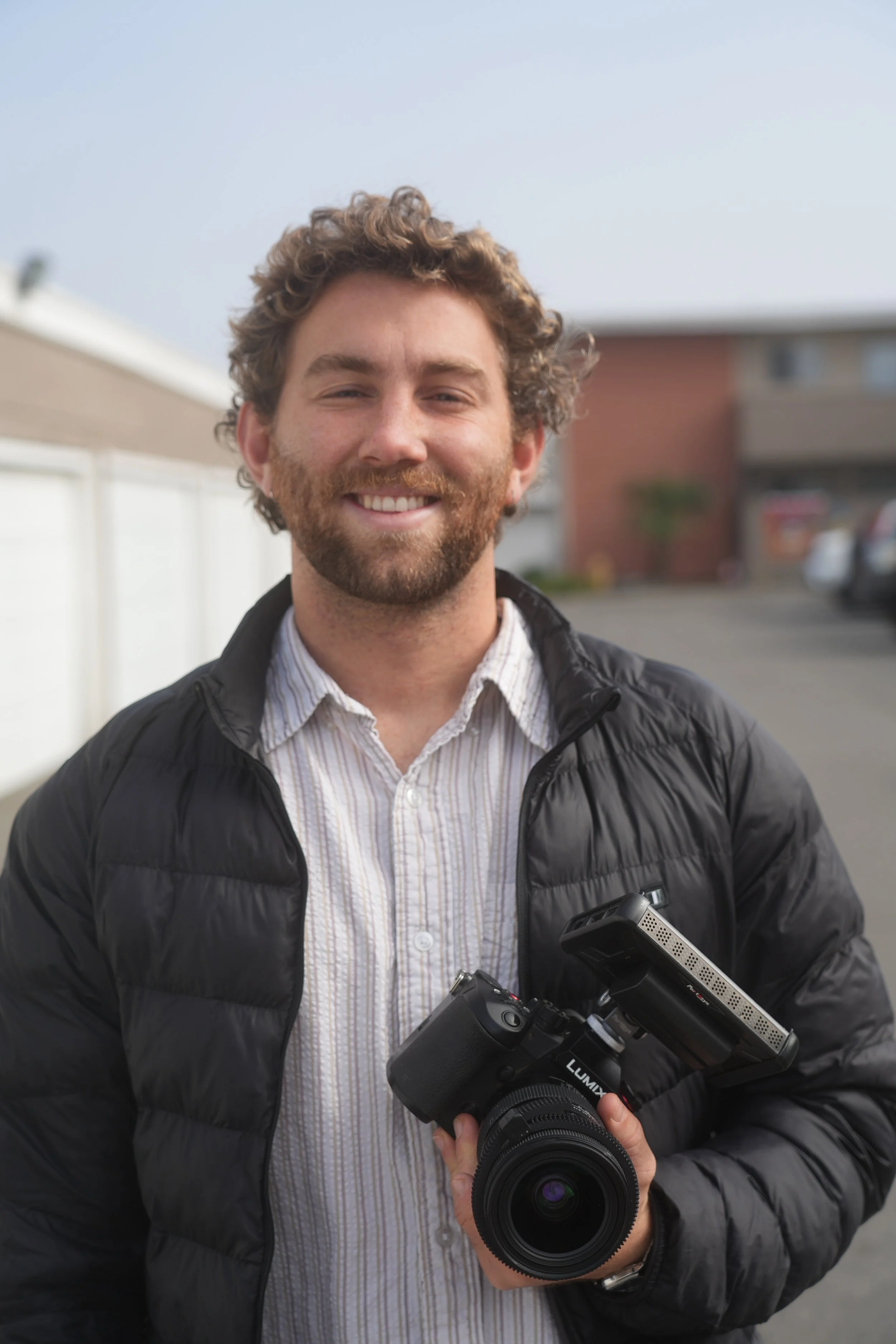 A man with curly hair, a beard, and a smile, holding a camera, standing outdoors in a parking lot with buildings in the background.