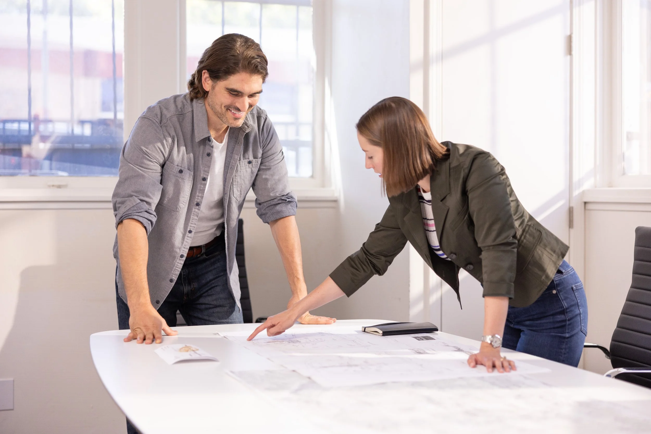 A man and a woman are leaning over a table, examining documents or plans together, smiling indoors with natural light from windows.