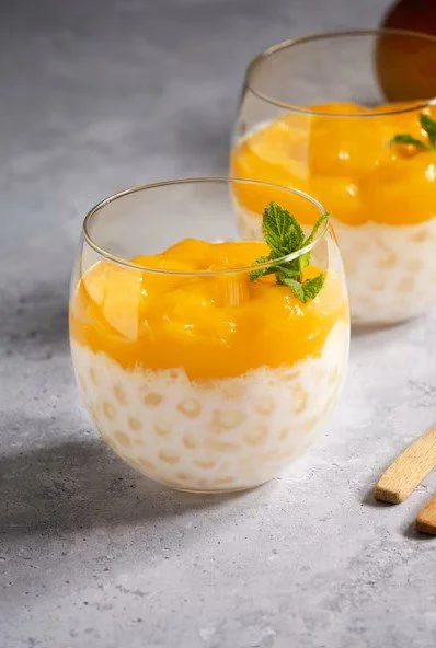 Two glasses of mango pudding topped with mango chunks and mint leaves, on a gray surface.