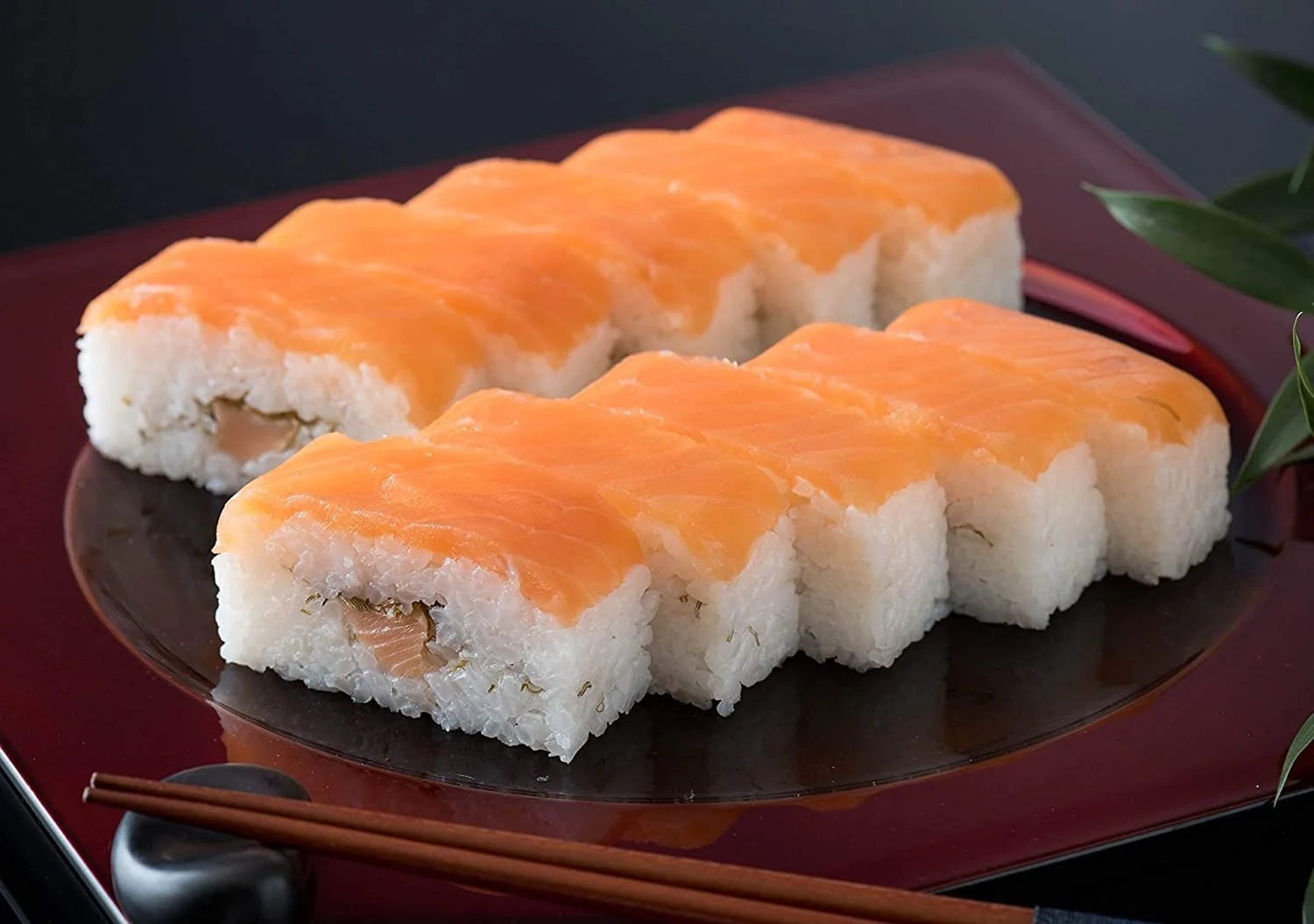 Sliced sushi roll topped with salmon, served on a black and red plate with a sprig of greenery and chopsticks.