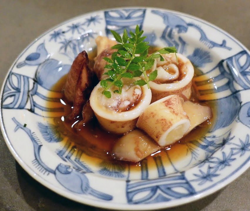 Sliced squid stuffed with filling, garnished with a sprig of green herbs, served with lemon wedge and a soy-based sauce in a blue and white decorative bowl.
