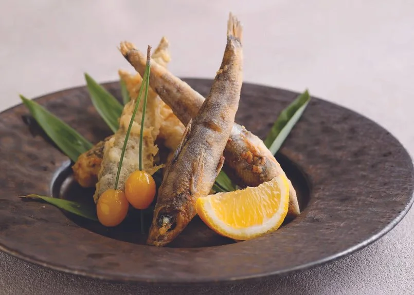 Fried small fish garnished with lemon wedge, green leaves, and cherries on a round dark plate.