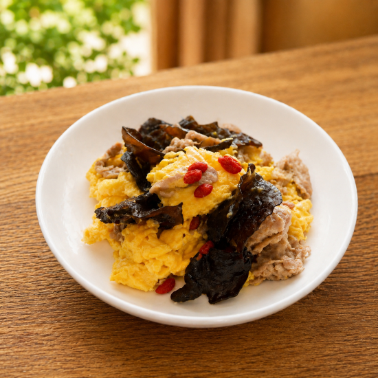 A bowl of mashed potatoes topped with fried pork skin, goji berries, and shredded pork on a wooden table.