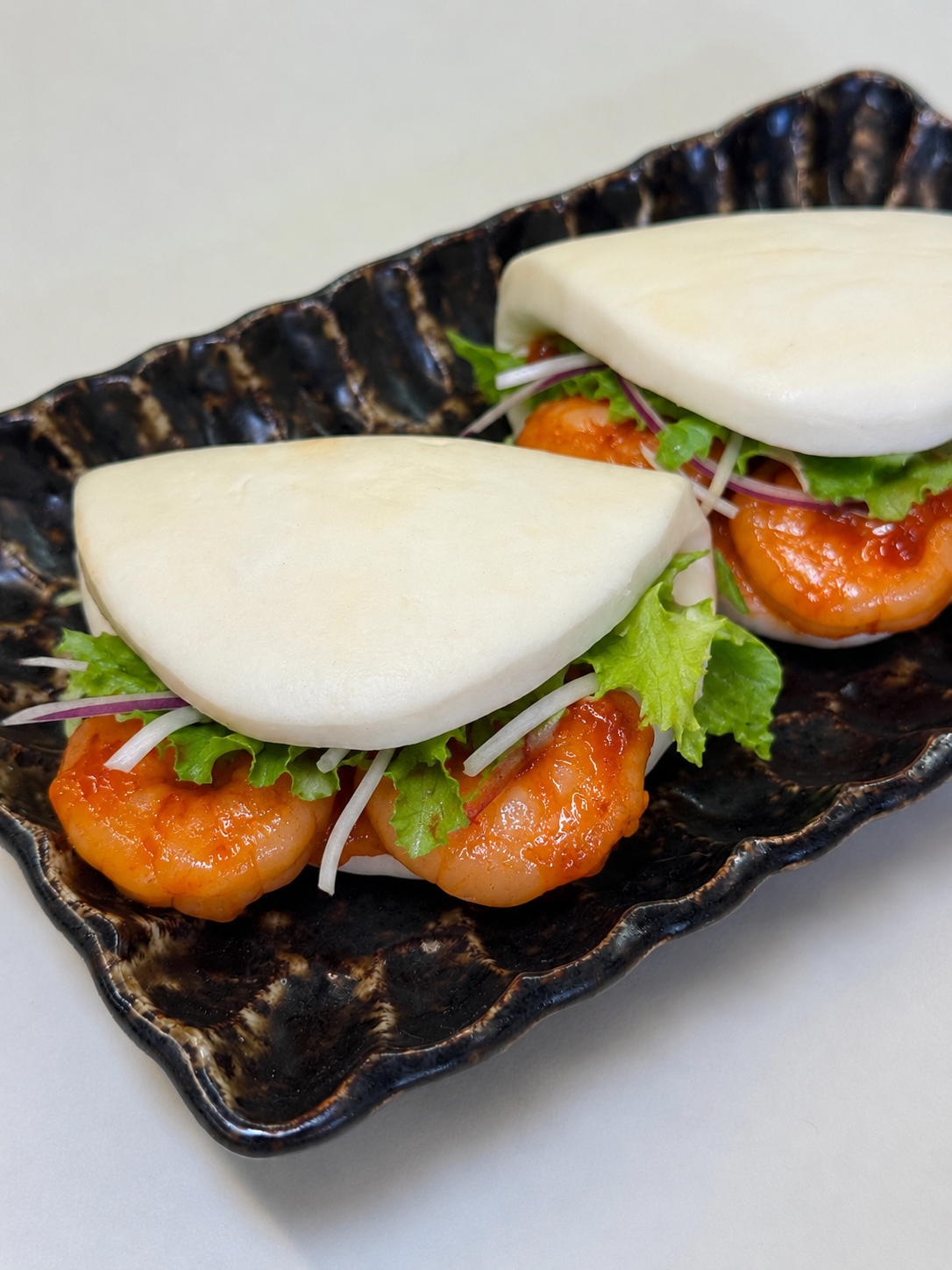 Two steamed bao buns filled with shrimp, lettuce, and microgreens on a black textured plate.