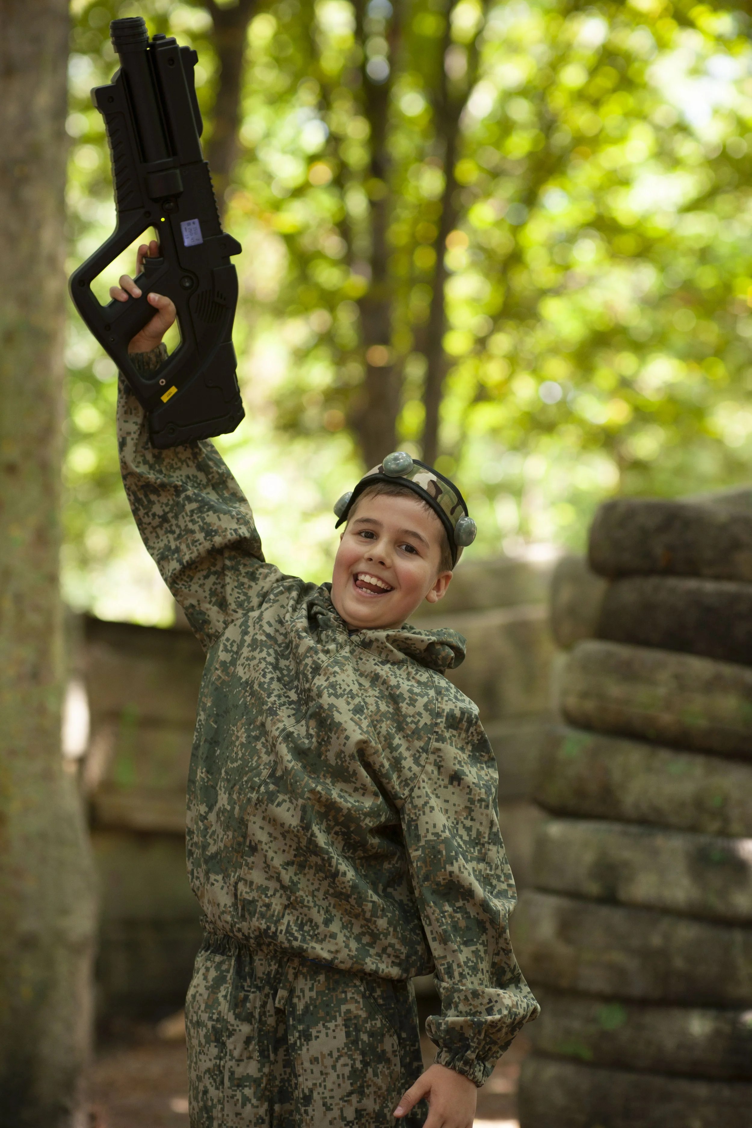 Young boy in camouflage clothes and goggles, smiling and holding a toy gun, in a wooded area.
